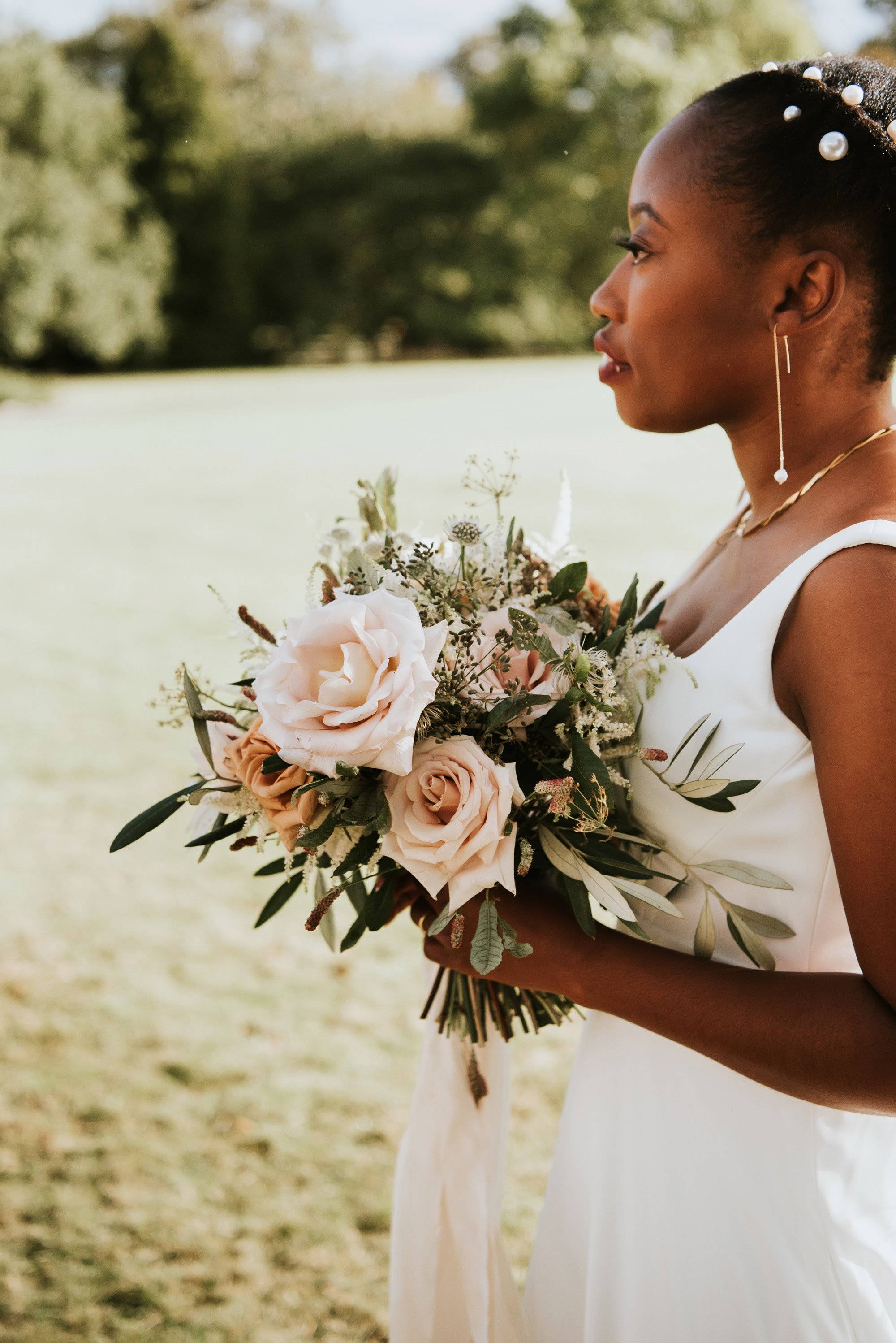Modern black bride wearing pearl drop earrings and pearls in her hair carrying bouquet of pale pink roses and greenery outside her stately home wedding venue