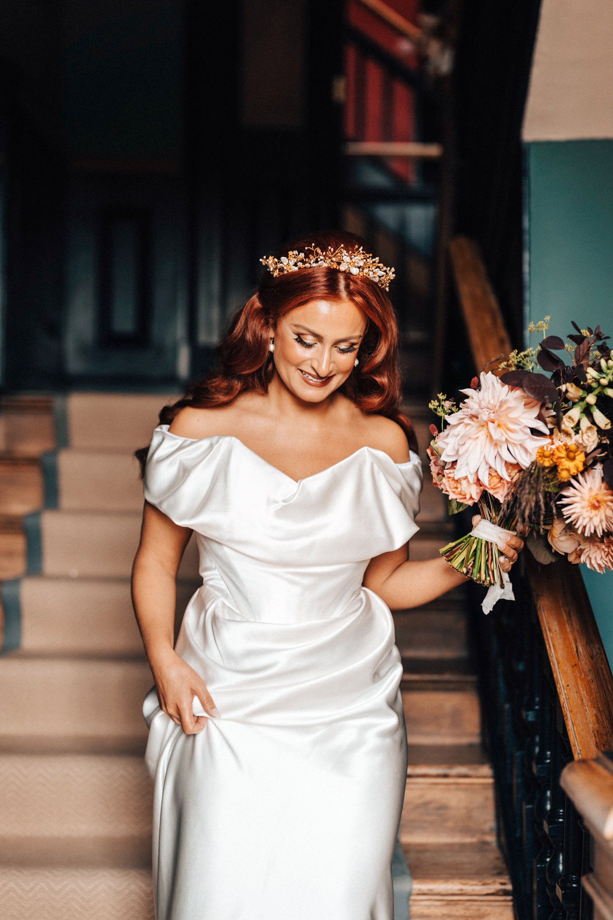 Modern bride with red hair wearing boho pearl tiara, pearl earrings and Alexander Mcqueen off shoulder wedding dress holding wild pink bouquet walking downstairs of stately home wedding venue