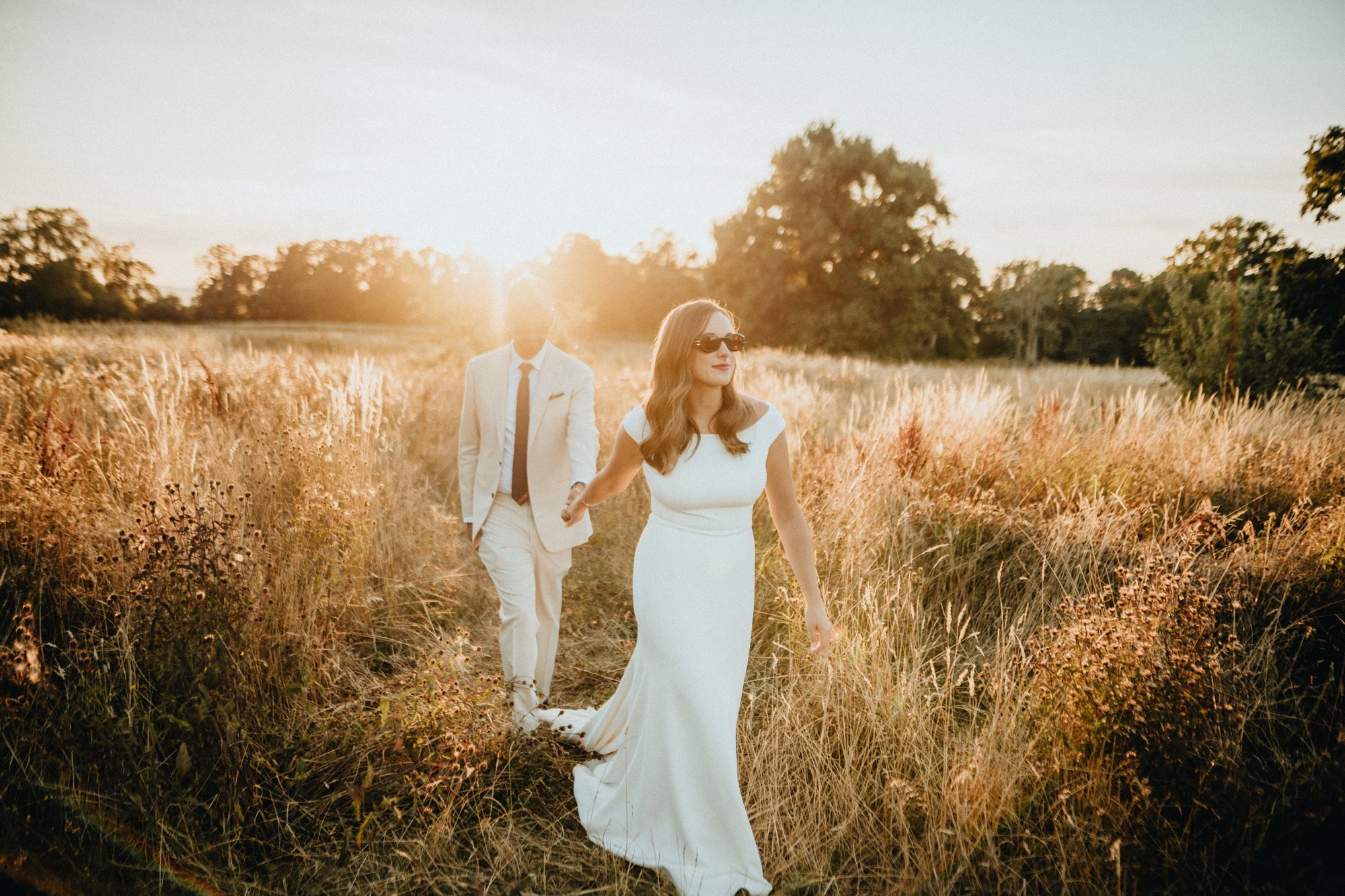 Cool bride and groom wearing sunglasses while walking through a meadow at sunset