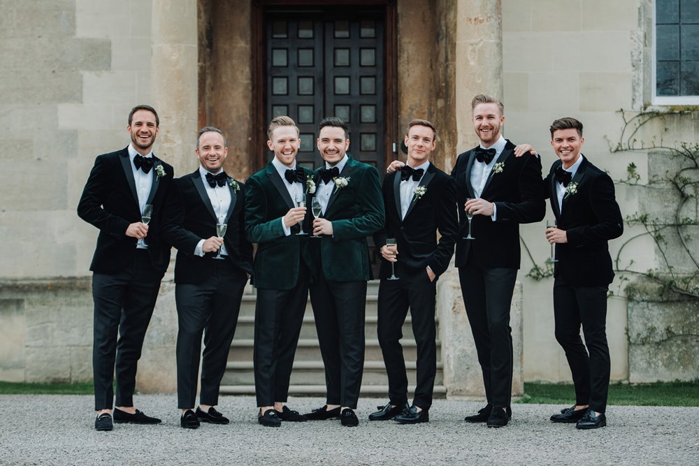 Gay wedding party: 2 grooms and their 5 groomsmen standing outside their stately home wedding venue elmore court wearing matching velvet tuxedos 