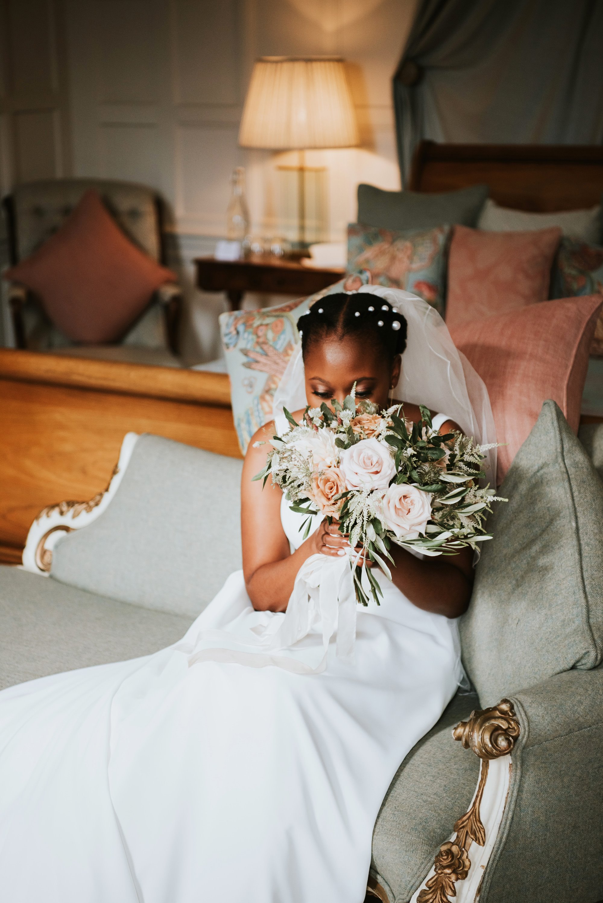 Bride sitting on chaise lounge in glamorous bridal suite of stately home pushes face into bouquet of blush pink and greenery