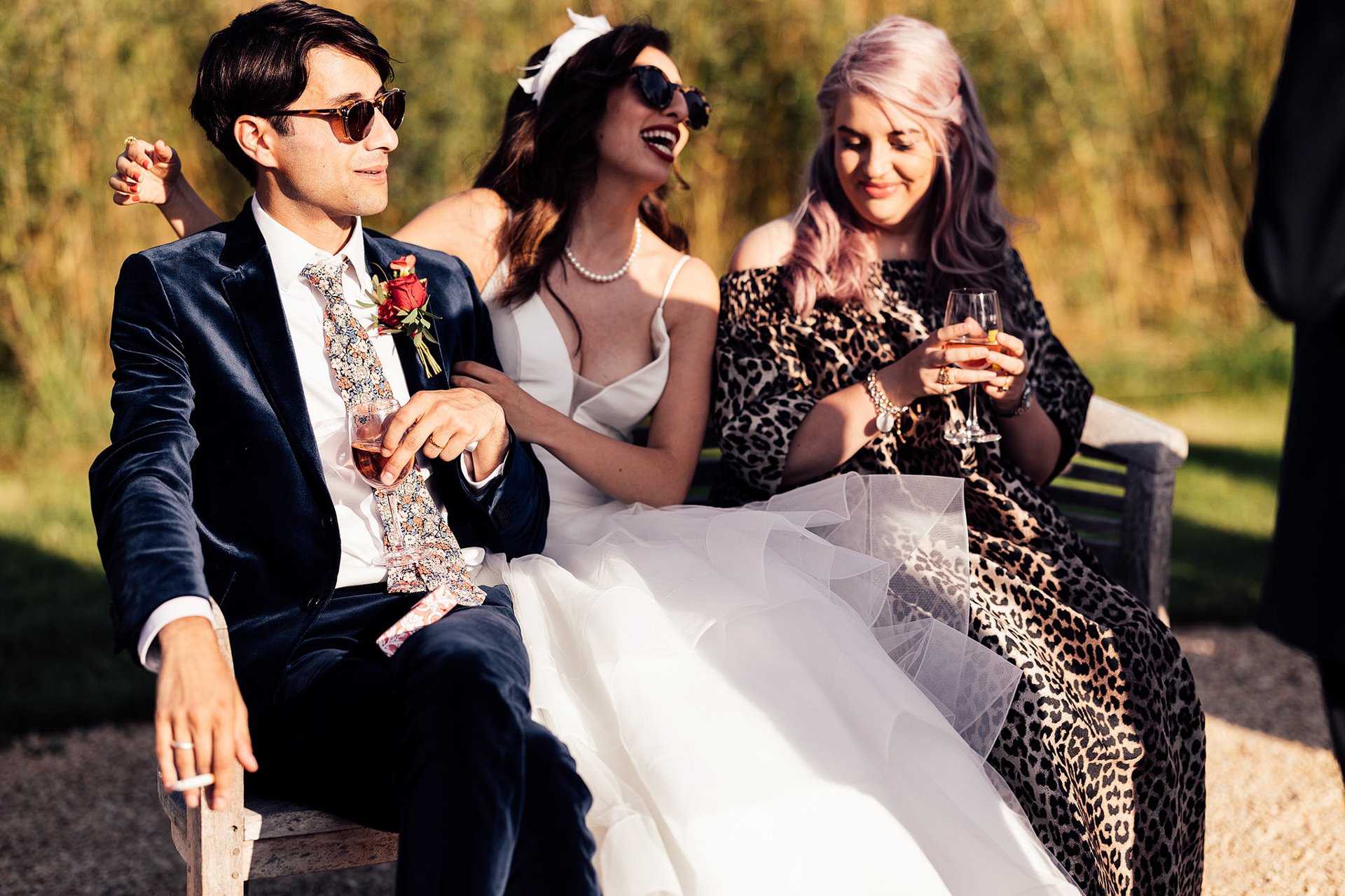 Beautiful bride and groom wearing sunglasses sit outside their wedding venue. Groom in blue velvet suit and bride in princess dress and designer headpiece