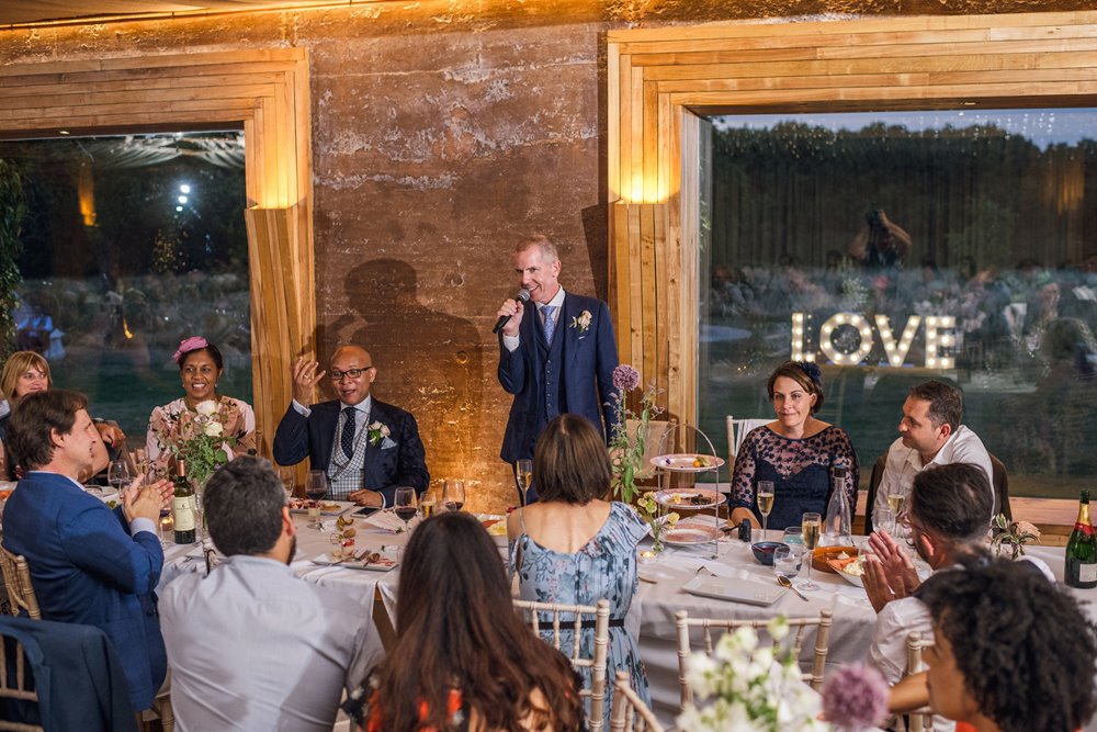 Gay wedding speeches, one groom makes speech whilst other listens inside rammed earth wedding venue with love light up letters seen through the window 