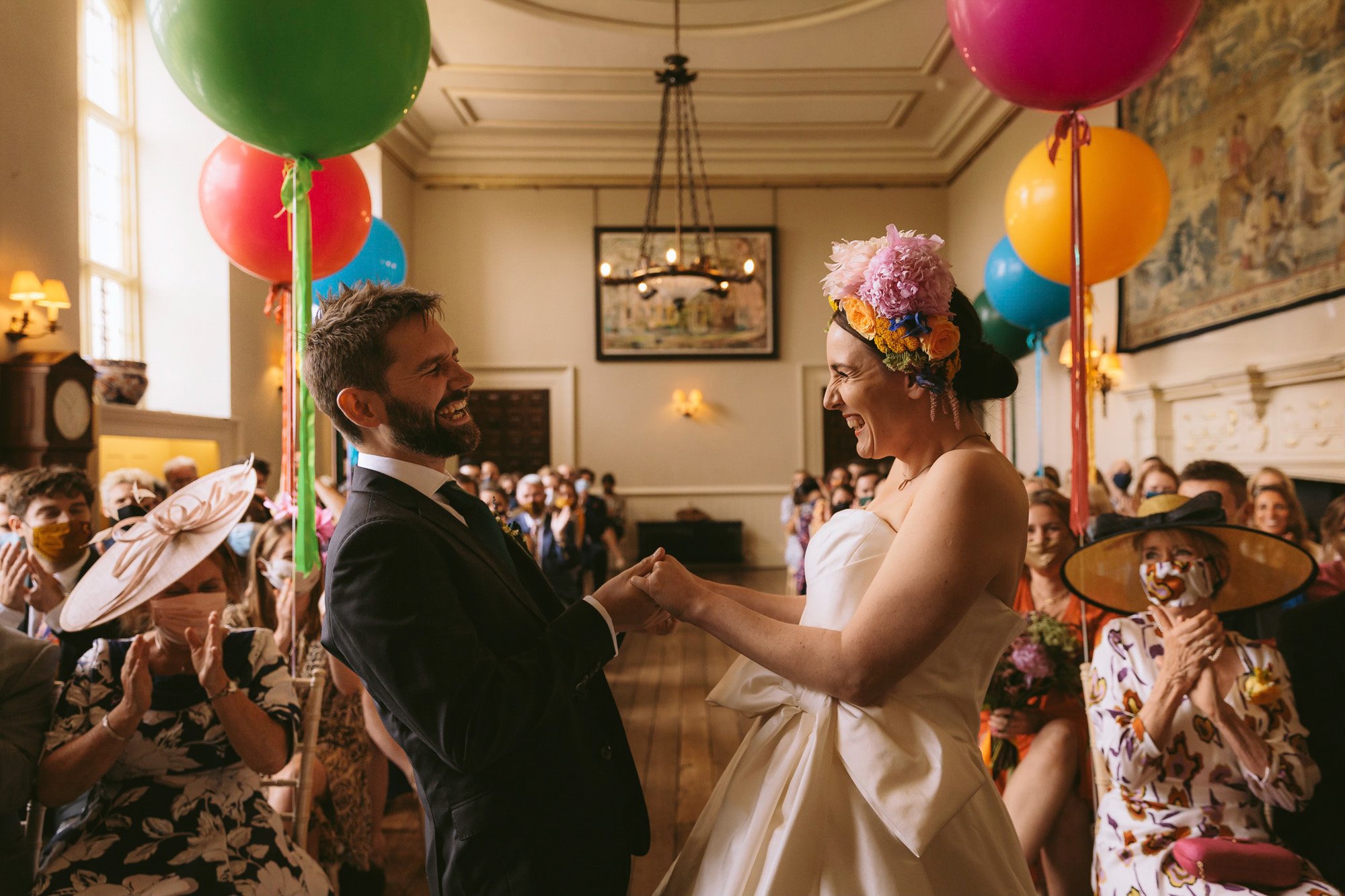 Festival bride in rainbow flower crown and groom hold hands and laugh in front of rainbow balloons and guests at their wedding ceremony in mansion house elmore court