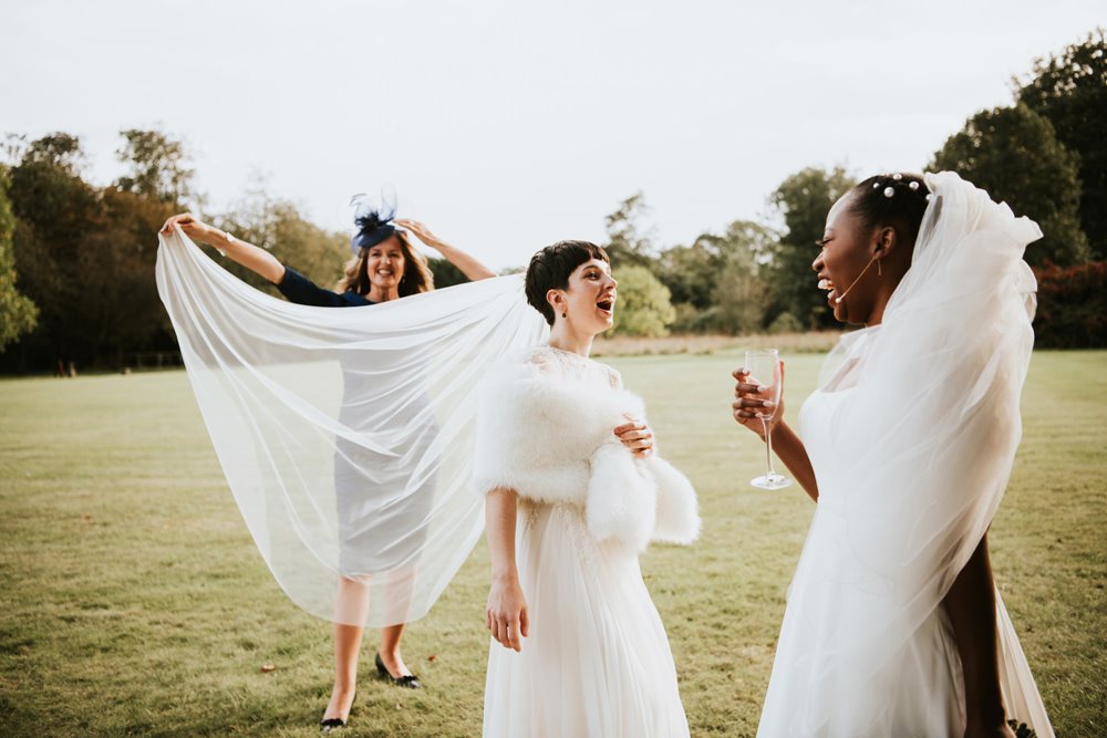 Two brides laugh and smile at each other at their Autumnal lesbian wedding holding drinks on the lawn whilst mother of bride holds out long veil in the wind