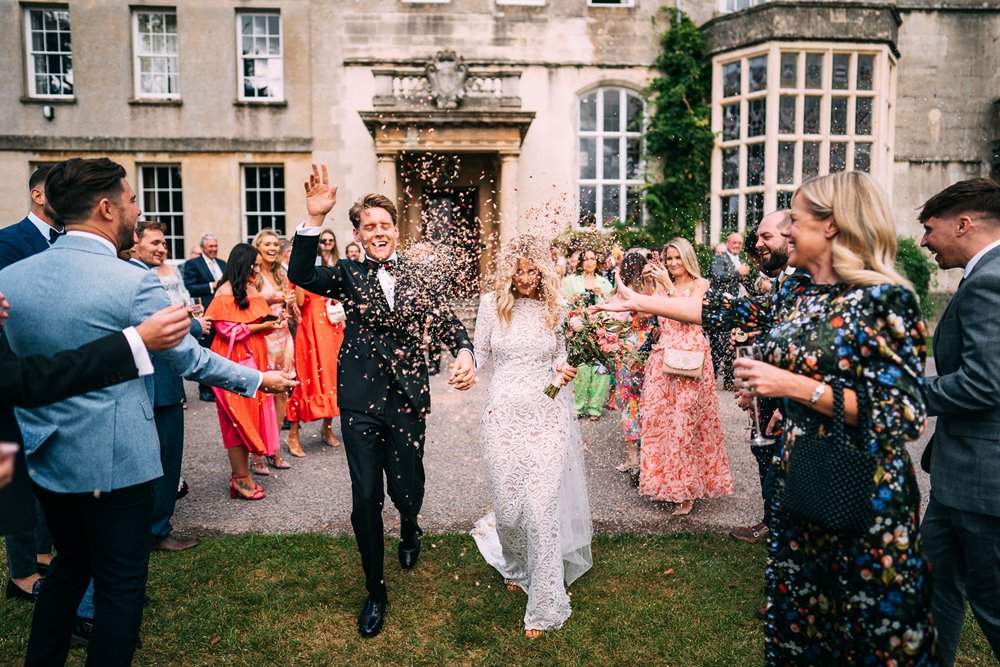 guests throwing colourful confetti on the newly wed couple as they walk outside over a beautiful stately home front lawn