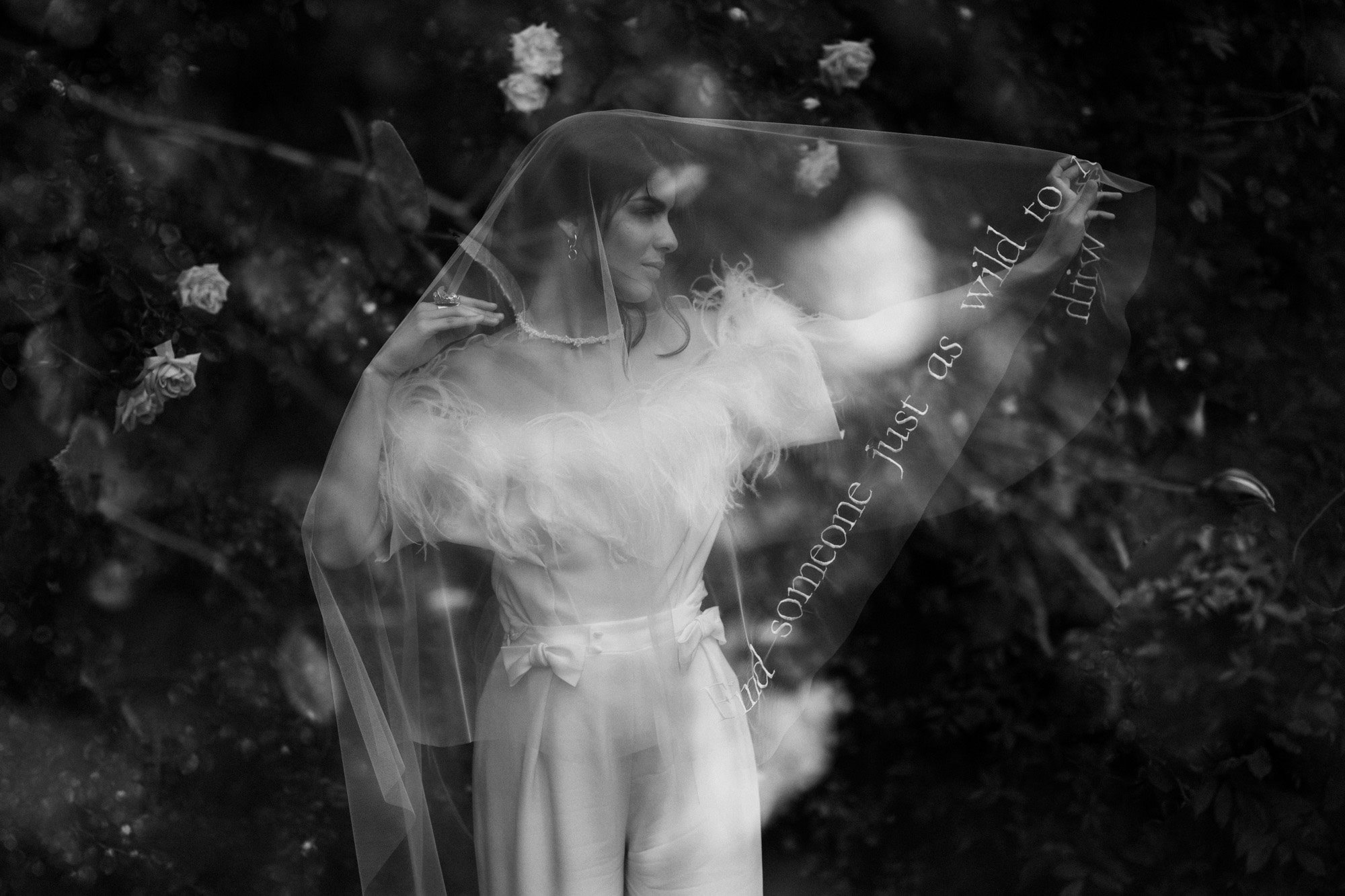 Modern chic wild bride underneath veil with writing wearing pearl necklace and feather jumpsuit in moody black and white shot