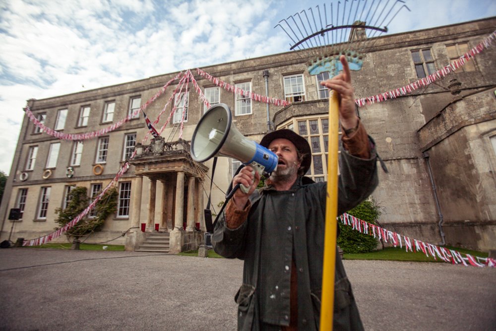 Hamish Guerrini the white rabbit of glastonbury stands with garden rake and megaphone in front of Secret garden party wedding venue mansion house elmore court in the cotswolds