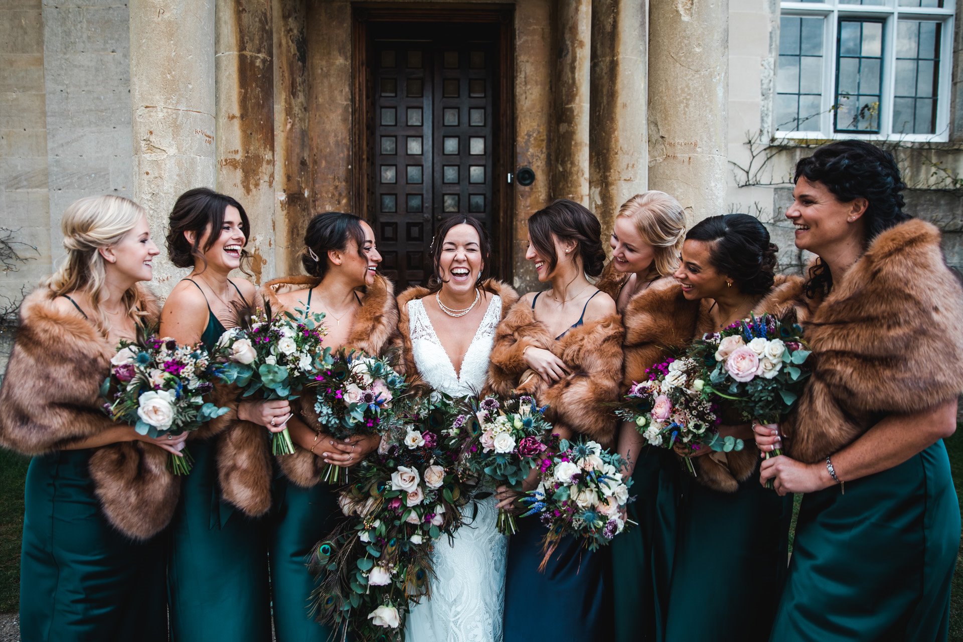 Big bridal party of 7 bridesmaids laugh and smile wearing faux fur in front of grade 2 listed exclusive use wedding venue