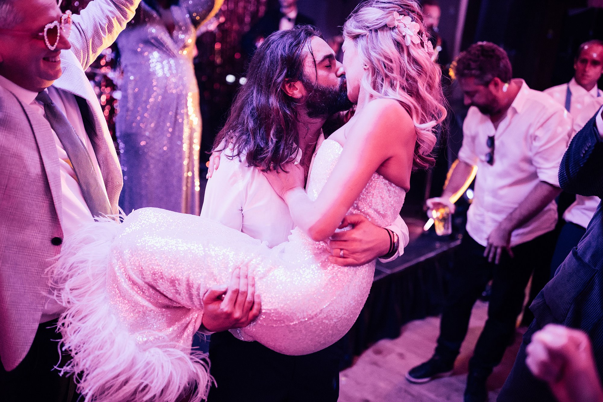 Groom carrying his bride while they kiss on the dance floor of their wedding reception in a soft pink glowing light