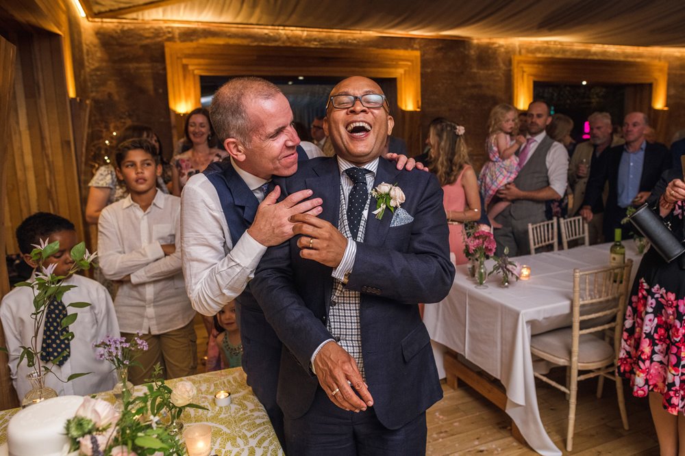 Gay couple hug and laugh as they cut cake at wedding reception in rammed earth venue with mud walls