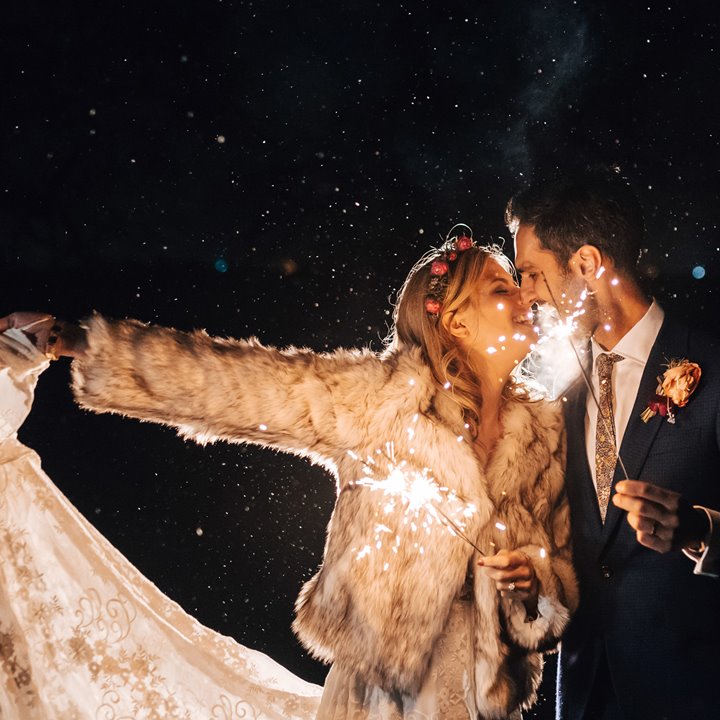 Boho bride in white lace wedding dress, white fur coat and flower crown holds her dress out to the side in one hand and a sparkler in the other as she leans in to kiss her modern groom in navy suit and patterned tie who is also holding sparkler at romantic modern boho wedding at night at wild wedding venue elmore court