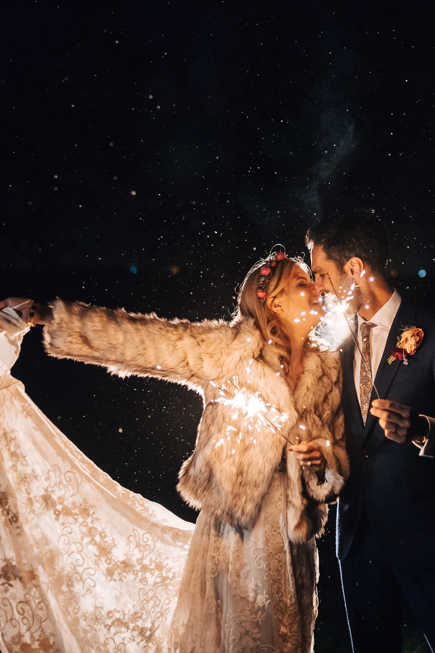 Boho bride in white lace wedding dress, white fur coat and flower crown holds her dress out to the side in one hand and a sparkler in the other as she leans in to kiss her modern groom in navy suit and patterned tie who is also holding sparkler at romantic modern boho wedding at night at wild wedding venue elmore court