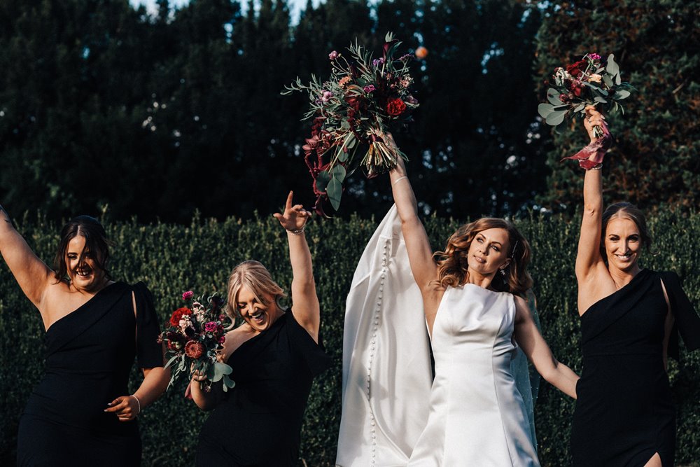 Cool bride and bridal party in black bridesmaids dresses holding up autumnal bouquets at dark and moody fun wedding in October