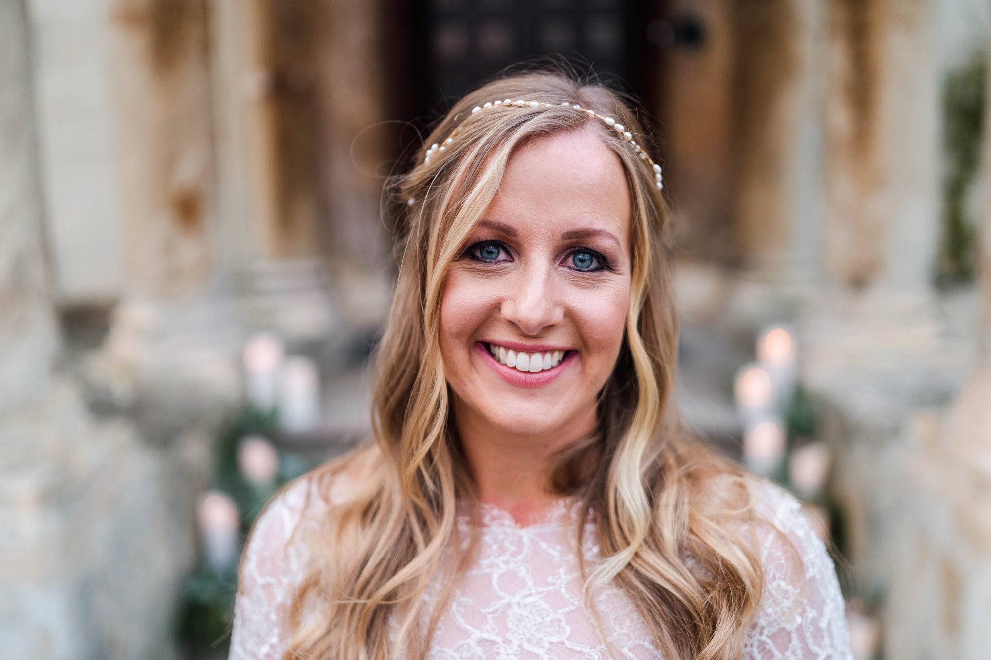 Blonde bride smiles at camera wearings simple pearl bridal headpiece and lace bodice dress on steps of stately home in cotswolds
