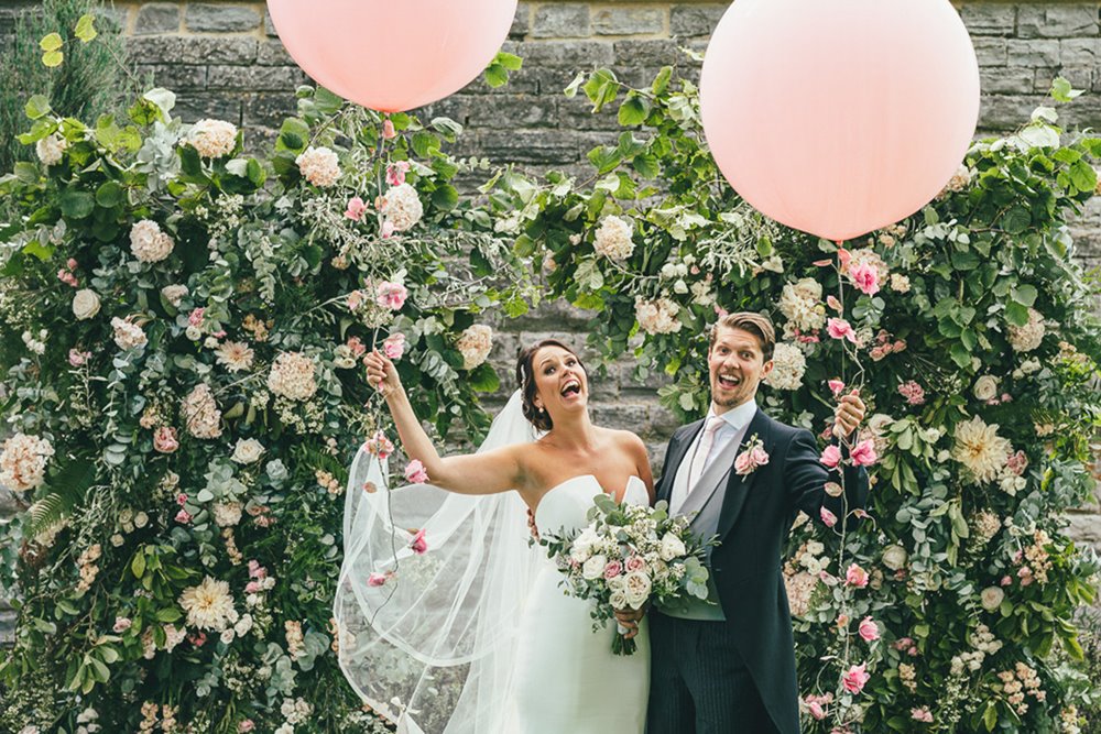 Fun newlywed couple pose with giant pink balloons in front of beautiful romantic wedding arch of pink and white roses