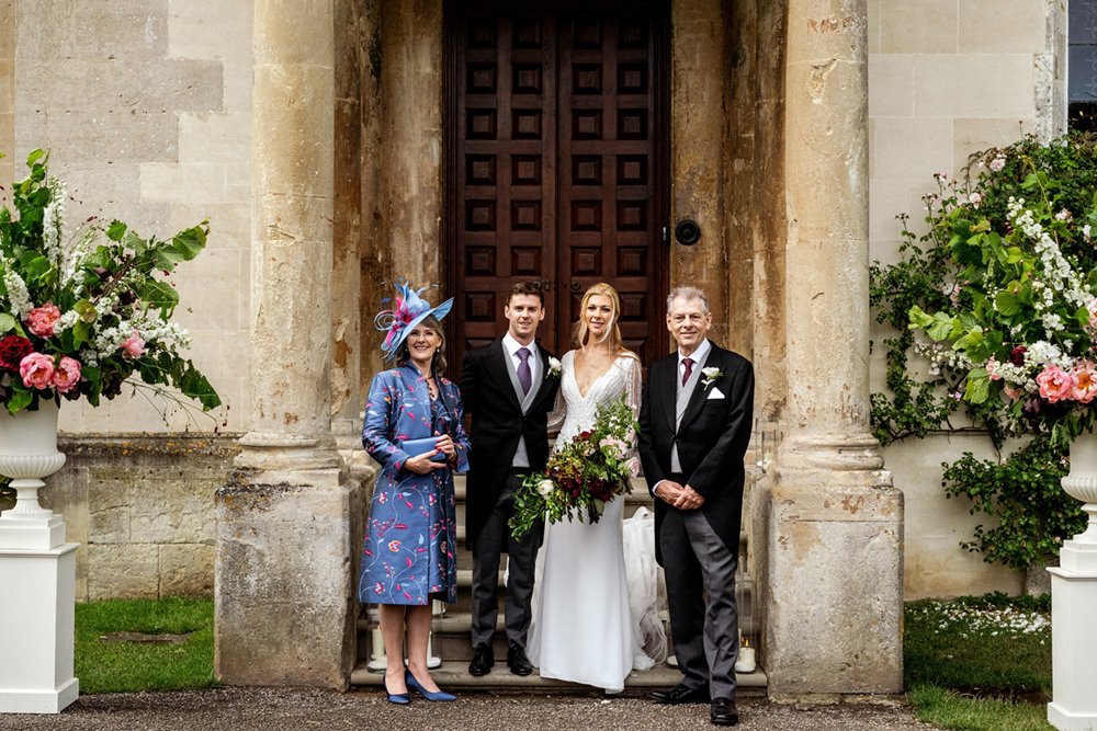 Bride and groom stand with family at entrance of stately home wedding venue elmore court. Mother of bride is wearing a beautiful unique dress coat in stunning purple fabric by Shibumi