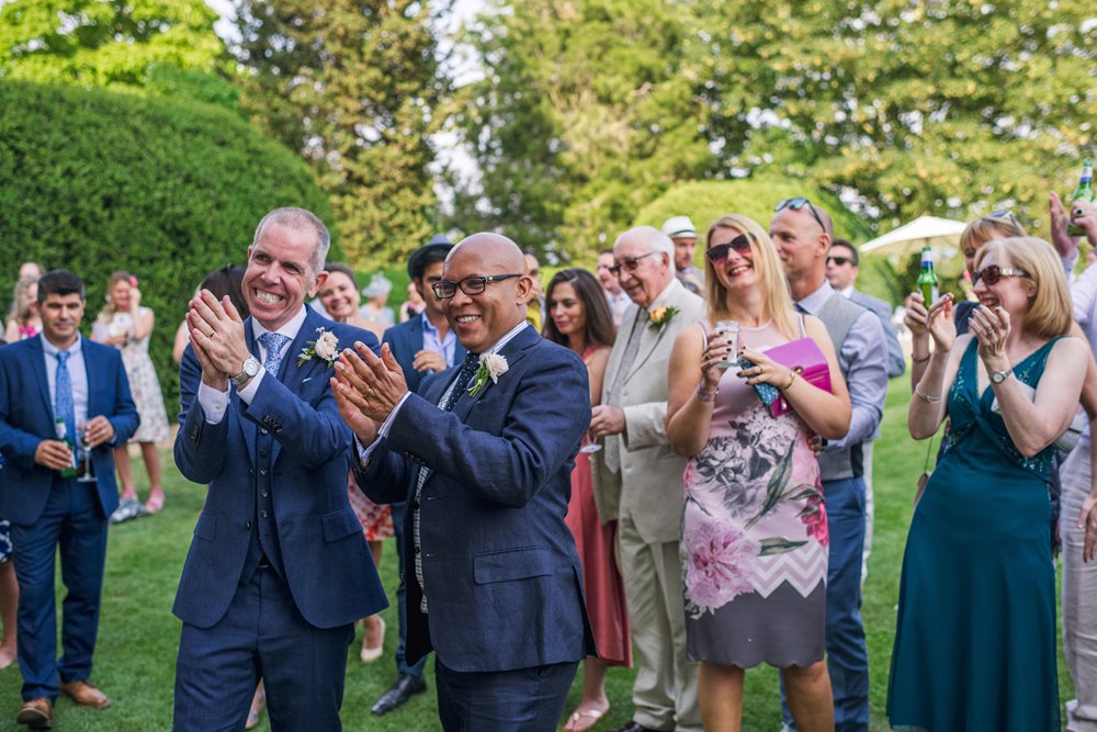 Two grooms clap and smile with their wedding guests at their garden party outdoor drinks reception in the costwolds 