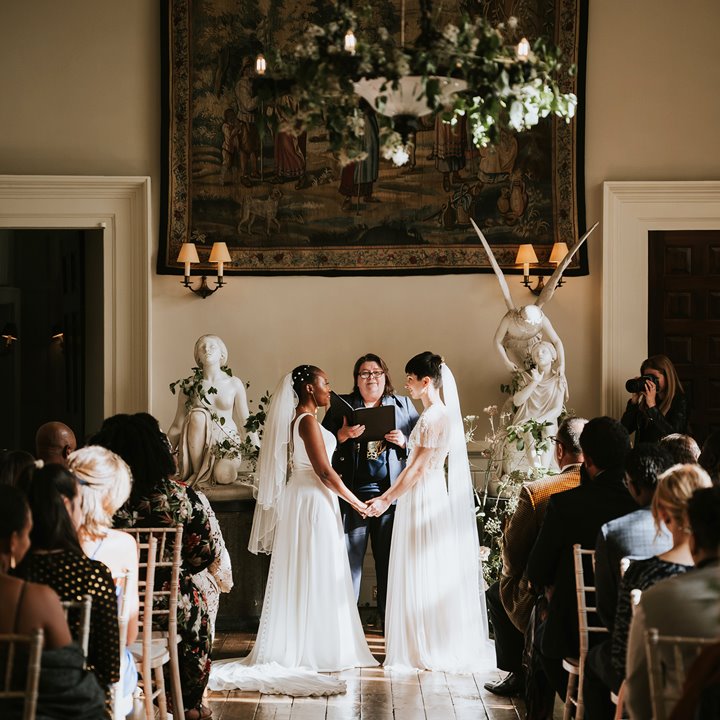 Two brides stand facing each other holding hands in their beautifully atmospheric wedding ceremony in the hall of stately home in the cotswolds