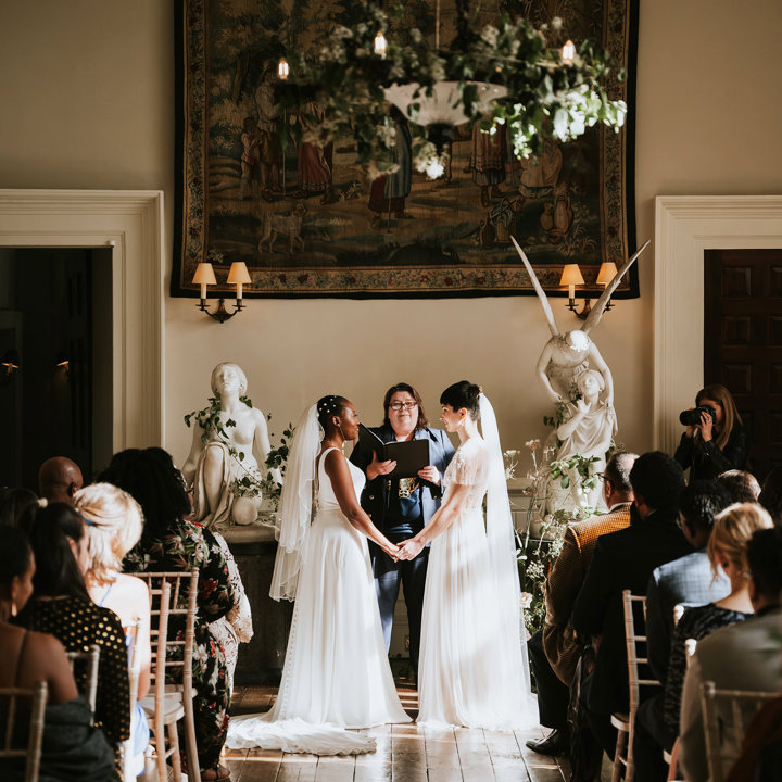 Two brides stand facing each other holding hands in their beautifully atmospheric wedding ceremony in the hall of stately home in the cotswolds