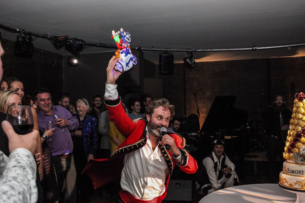 Hamish Guerrini the white rabbit of glastonbury in red coat holding toy car flying through air at massive party at mansion house elmore court