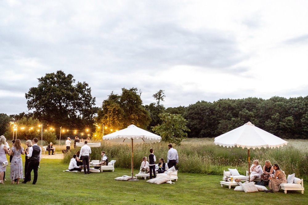 High end Boho luxe wedding reception outdoors in The Gillyflower meadow. Guests recline on low level seating under giant white parasols and chat around the fire pit underneath festoon lighting at a luxury Cotswolds wedding