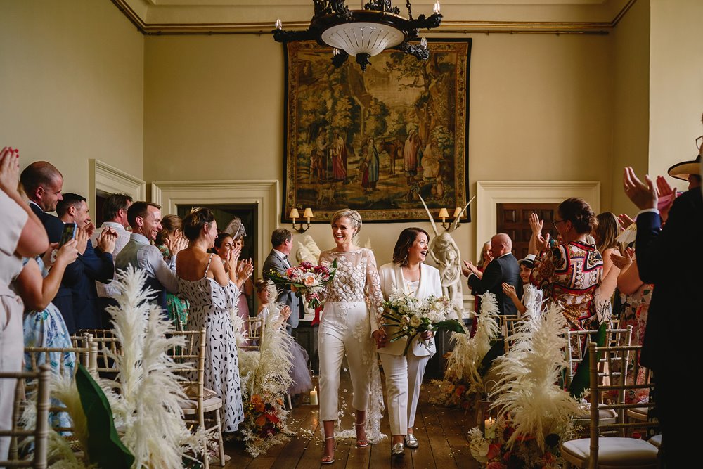 Lesbian wedding couple walk down the aisle smiling and holding hands - happily married at Elmore Court in Gloucestershire