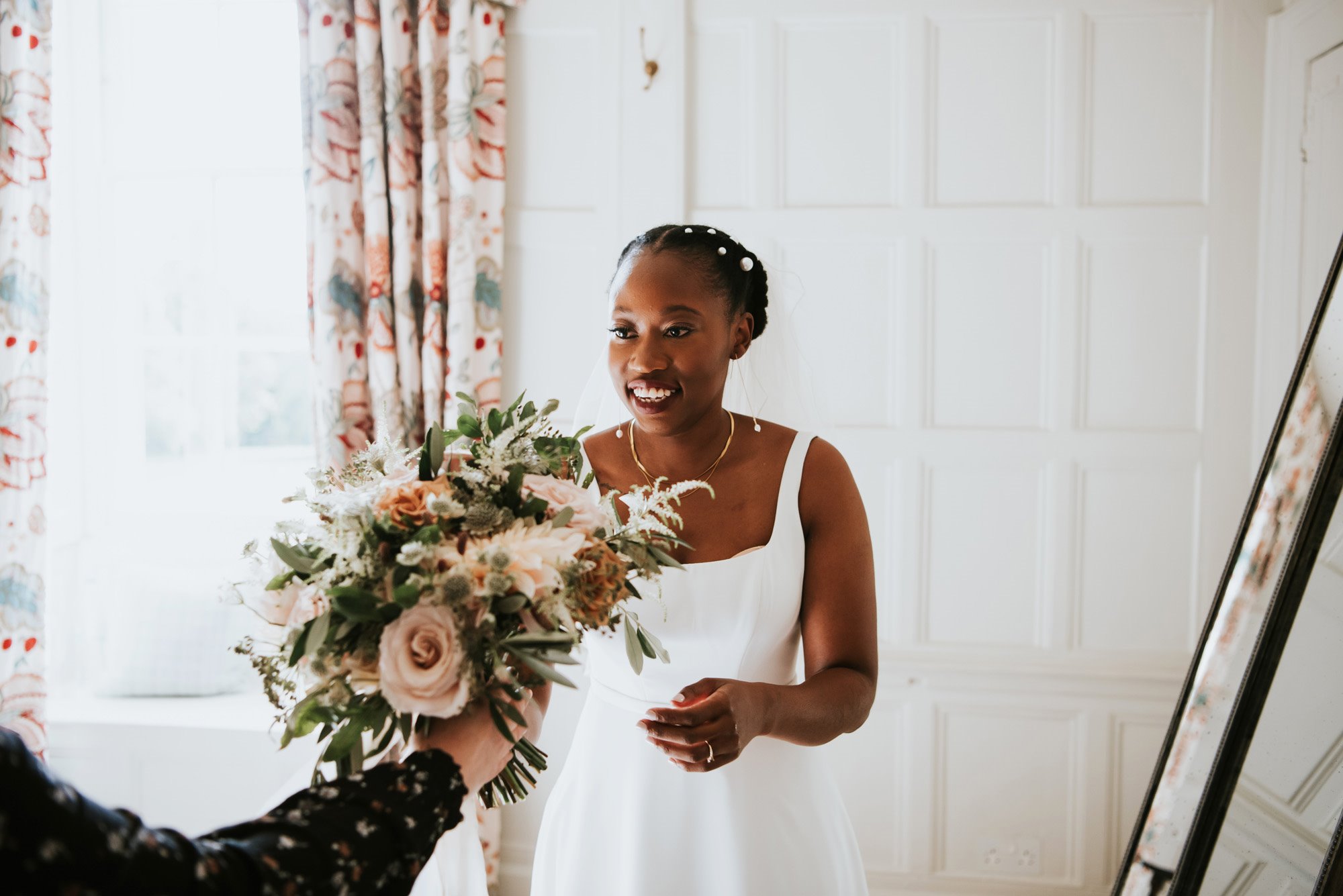 Bride with pearls in her afro hair in elegant updo gets handed her beautiful bouquet in luxurious bedroom of manor house venue