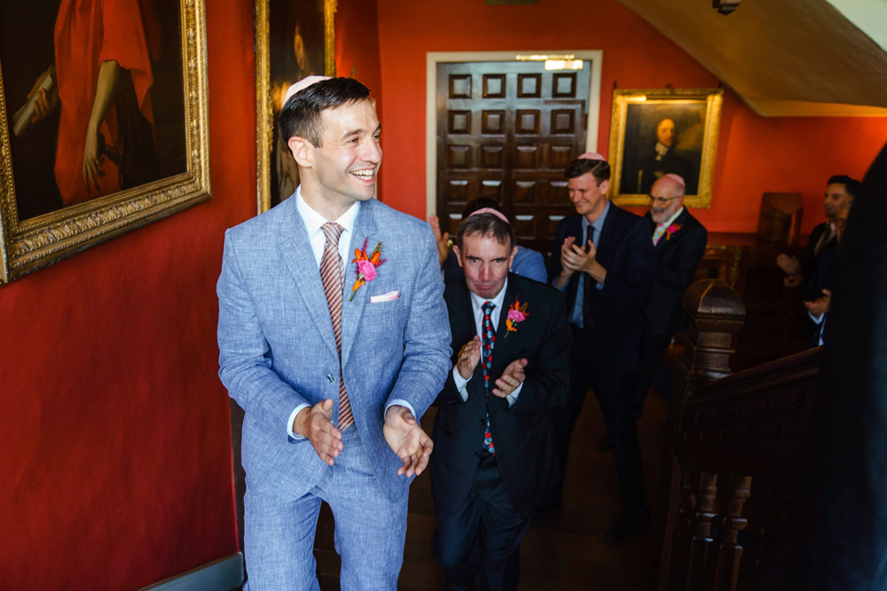 Jewish groom in bedeken procession clapping in a grey suit and pink kippah