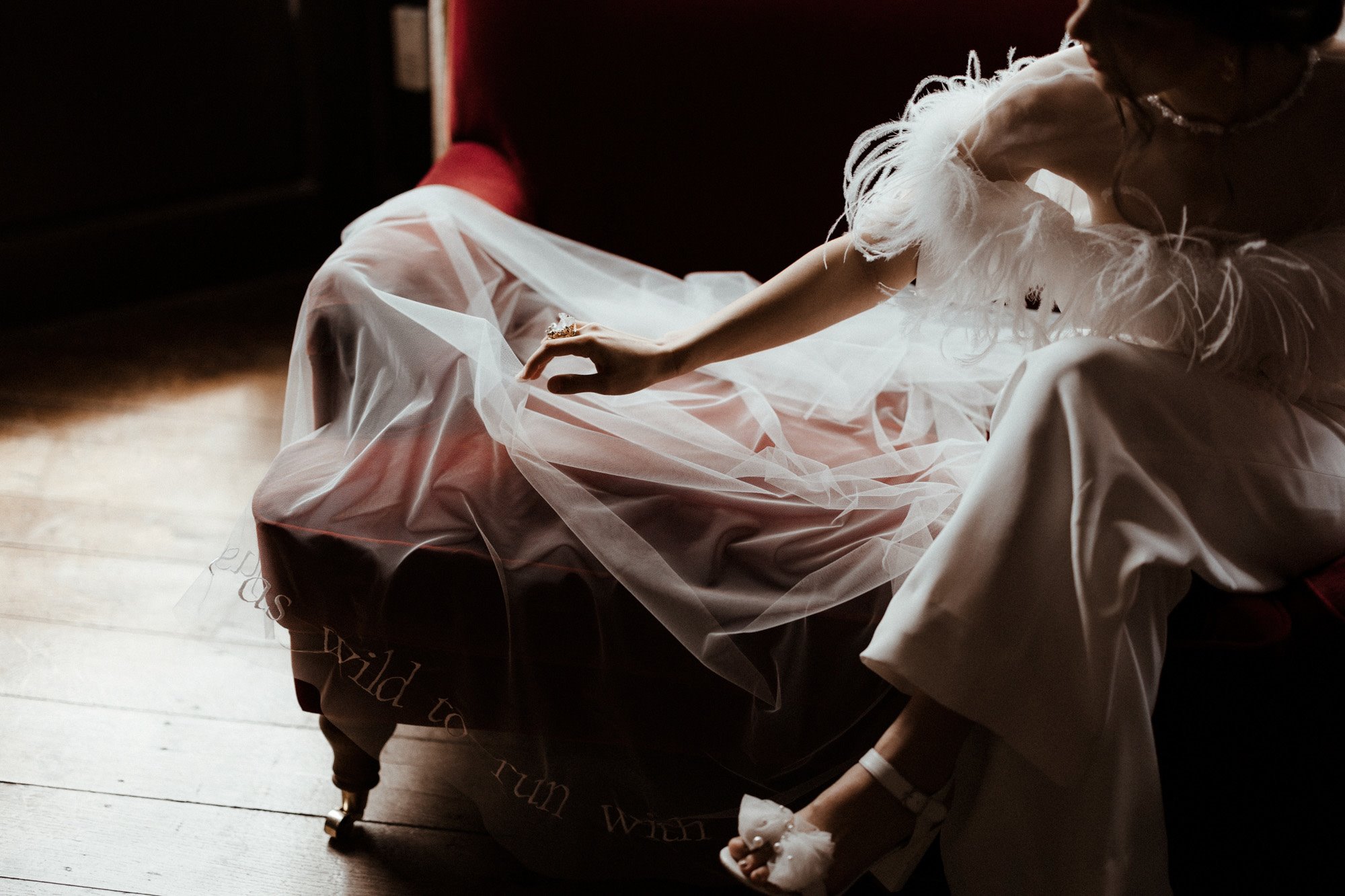 Chic modern bride wears Sexy feather jumpsuit, white shoes pearl with bow and pearl and pearl necklace on red velvet sofa at elmore court