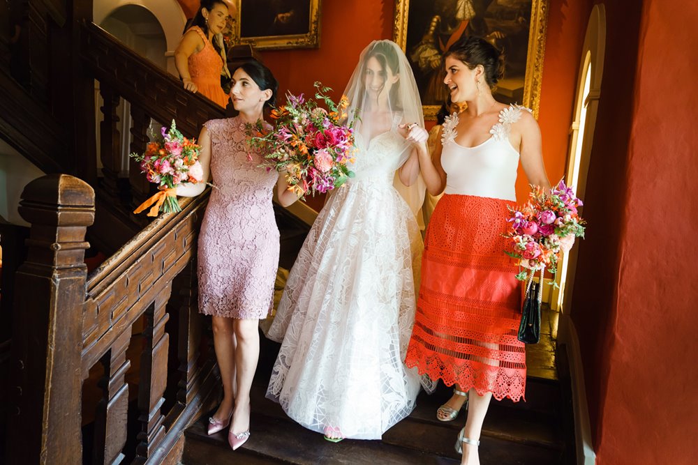 Bride in veil walks downstairs of Elmore Court holding bright pinkand orange bouquets 