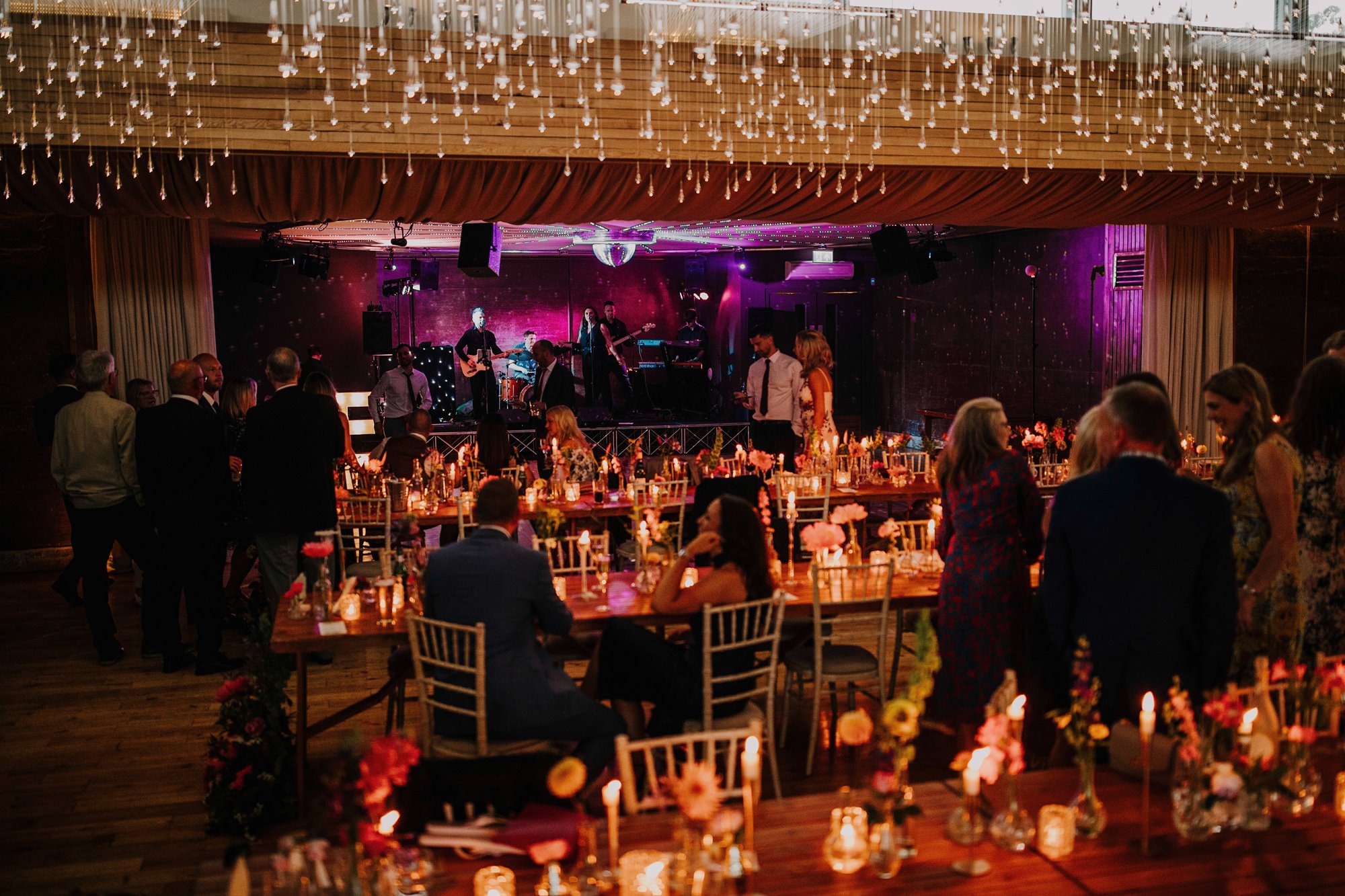 cosy wedding reception with flowers and candles flittering on tables while a band performs with a pink glowing lighting effects