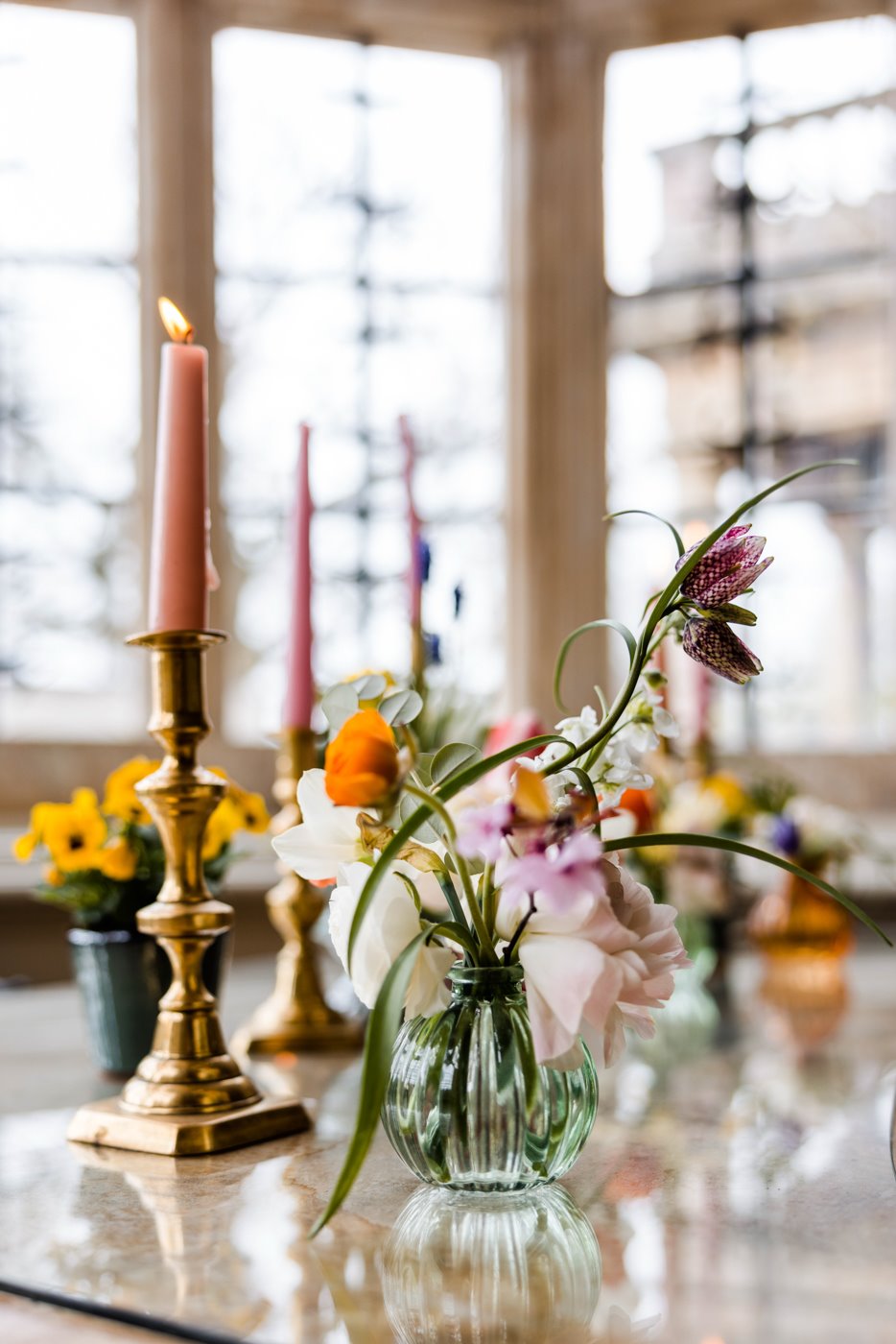 Delicate wild flowers in tiny glass vase and potted flowers with pink candlesticks in gold candlestick holder in mullioned glass windows of stately home wedding venue elmore court for a wild wedding fair
