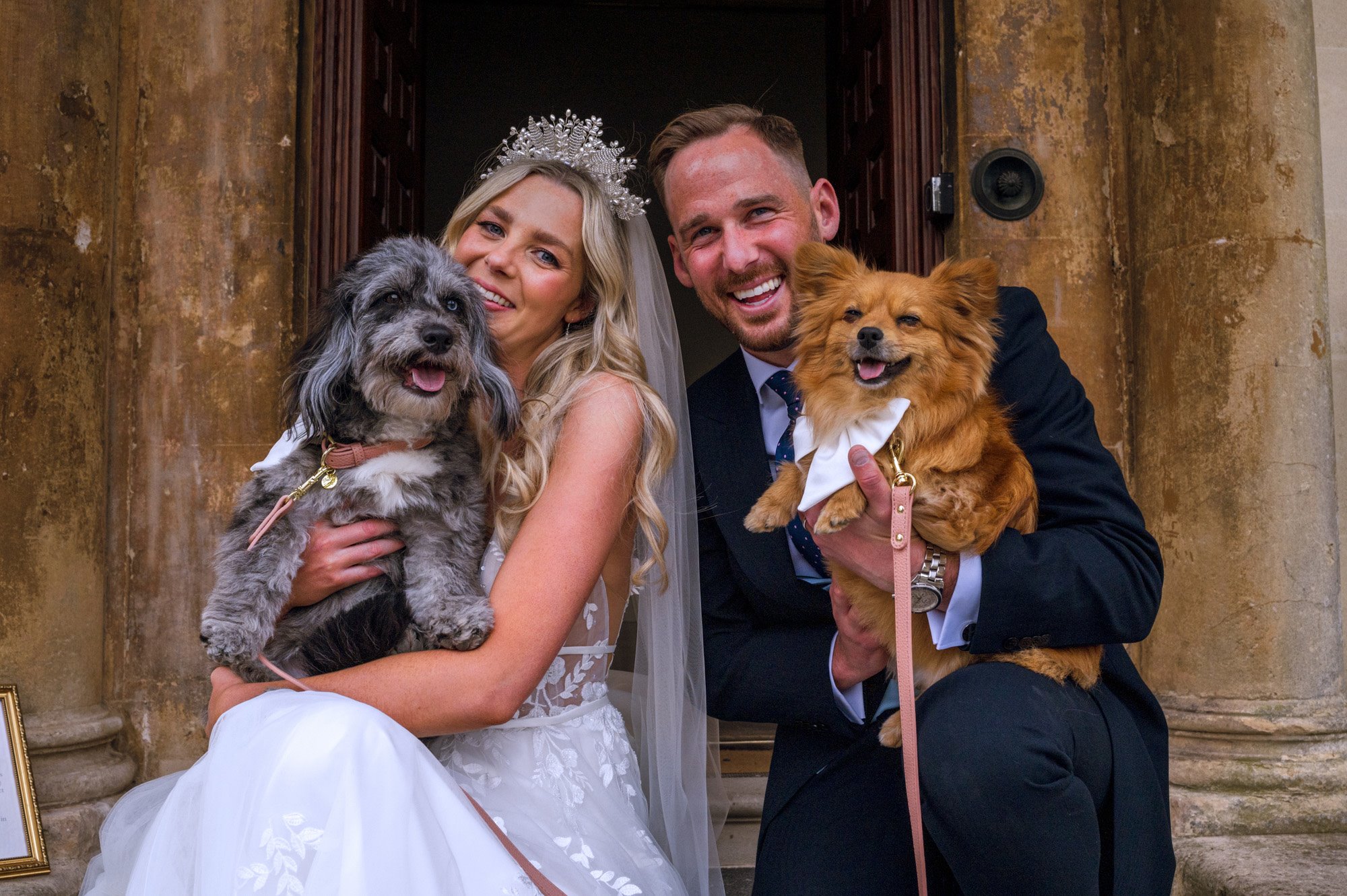 Boho bride and groom sit on steps of stately home wedding venue with their dogs in white bows. Bride wears boho lace and tulle wedding dress and pearl crown