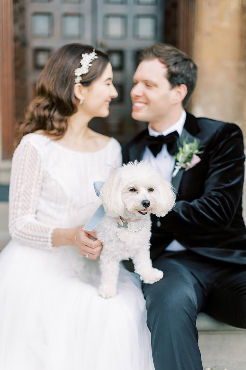 Newly wed could sitting on the steps of a lovely stately home with their furry dog friend on their laps