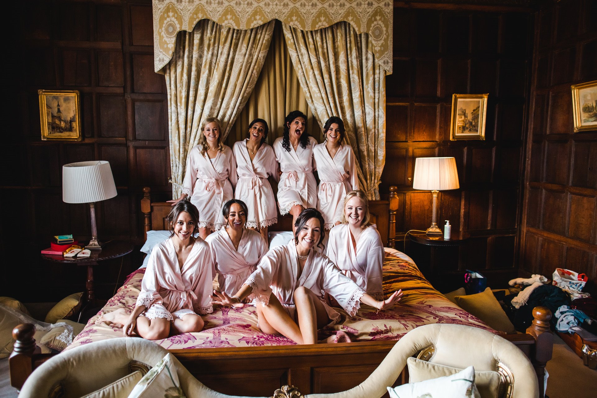 Big Bridal party of seven bridesmaids sit on huge bed in personalised dressing gowns on the morning of day 2 of weekend long wedding