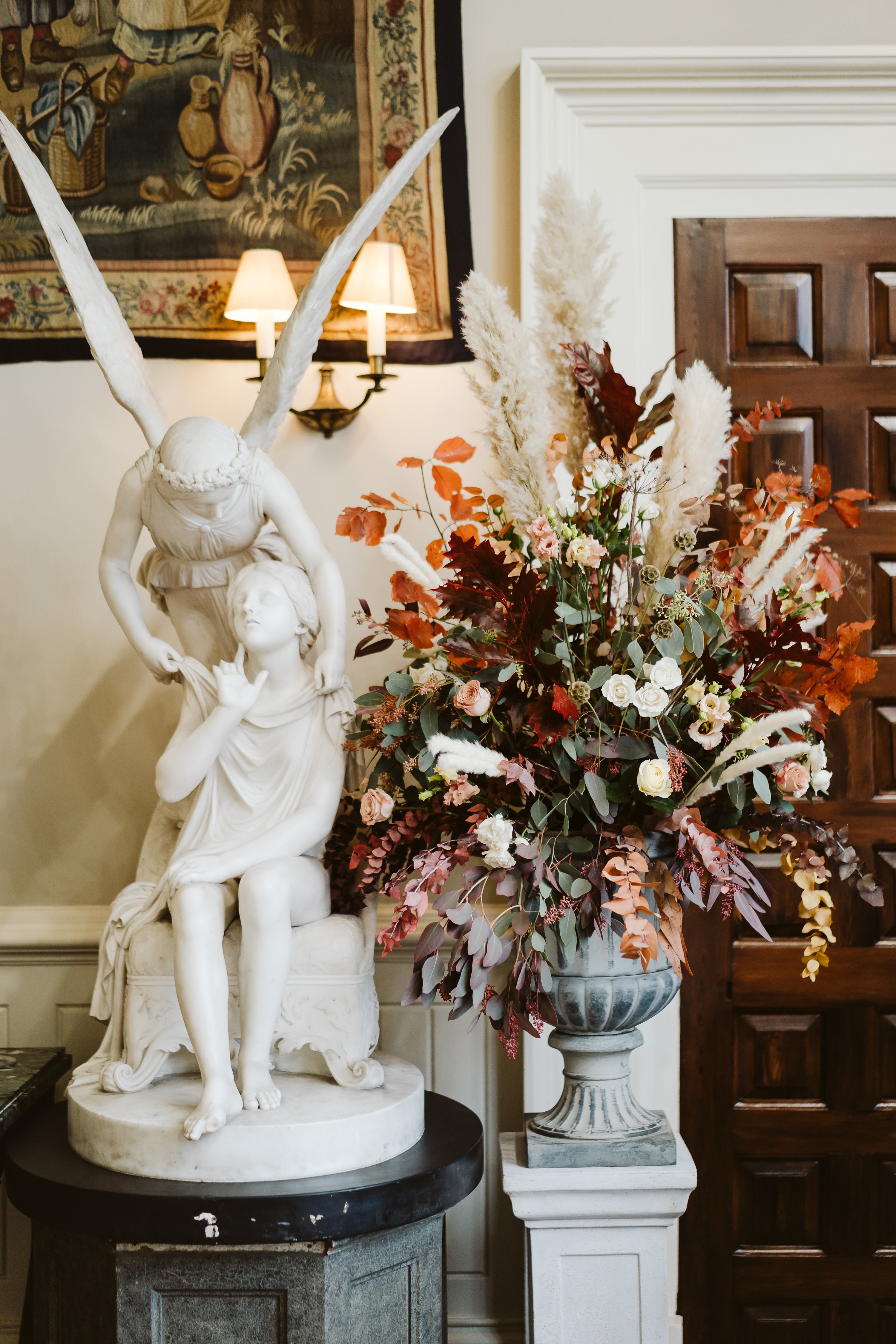 Autumn wedding at Elmore court with beautiful rustic floral arrangement in reds and oranges next to angel statue in the hall