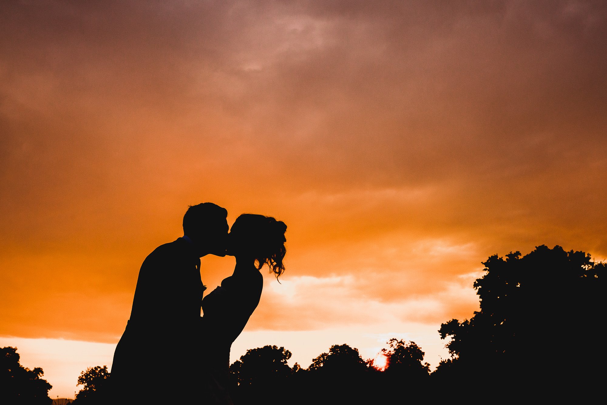 Silhouette of Bride and groom kiss against sunset sky at Elmore Court stately home in Gloucestershire