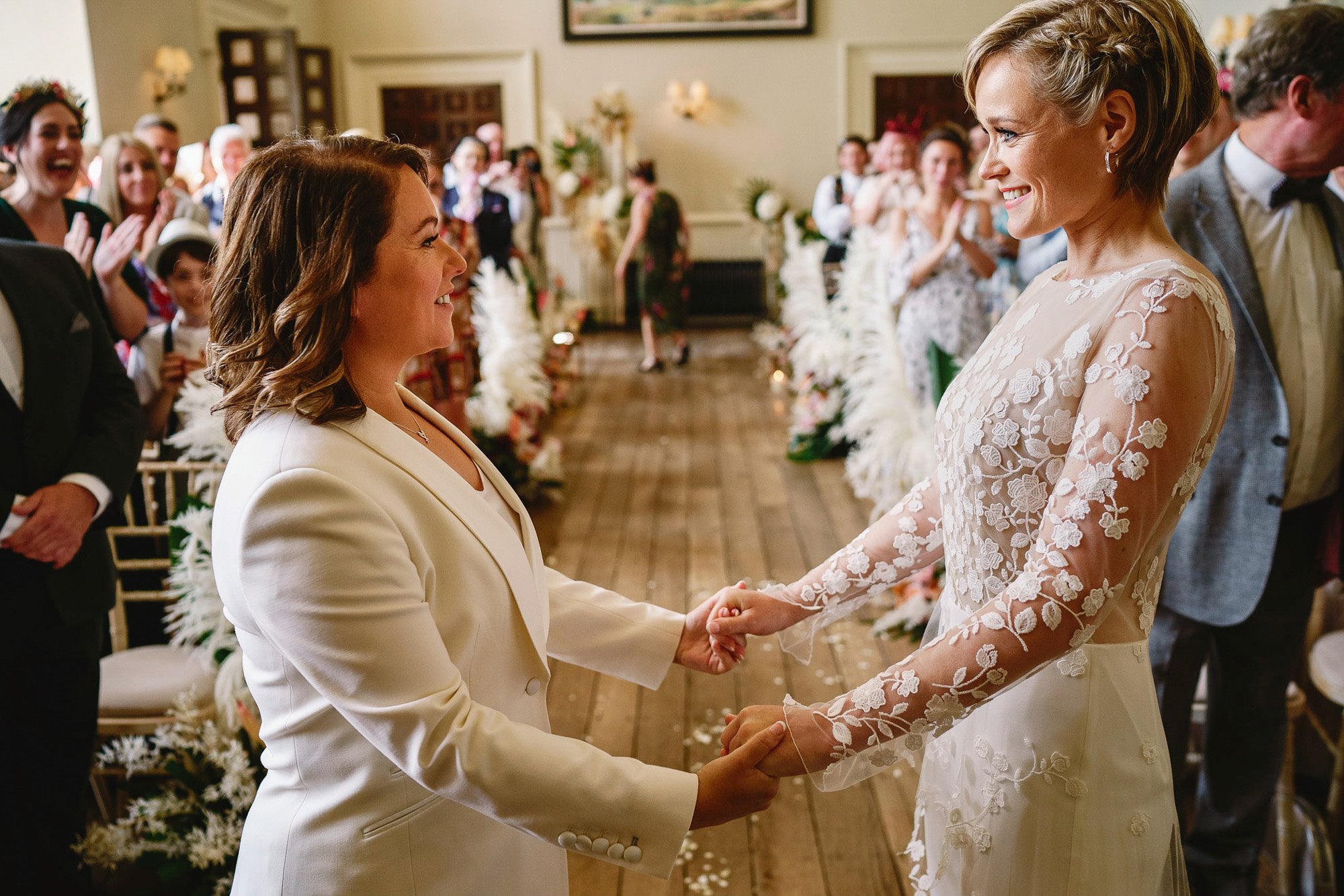 Two brides hold hands and smile at each other at their same sex wedding ceremony