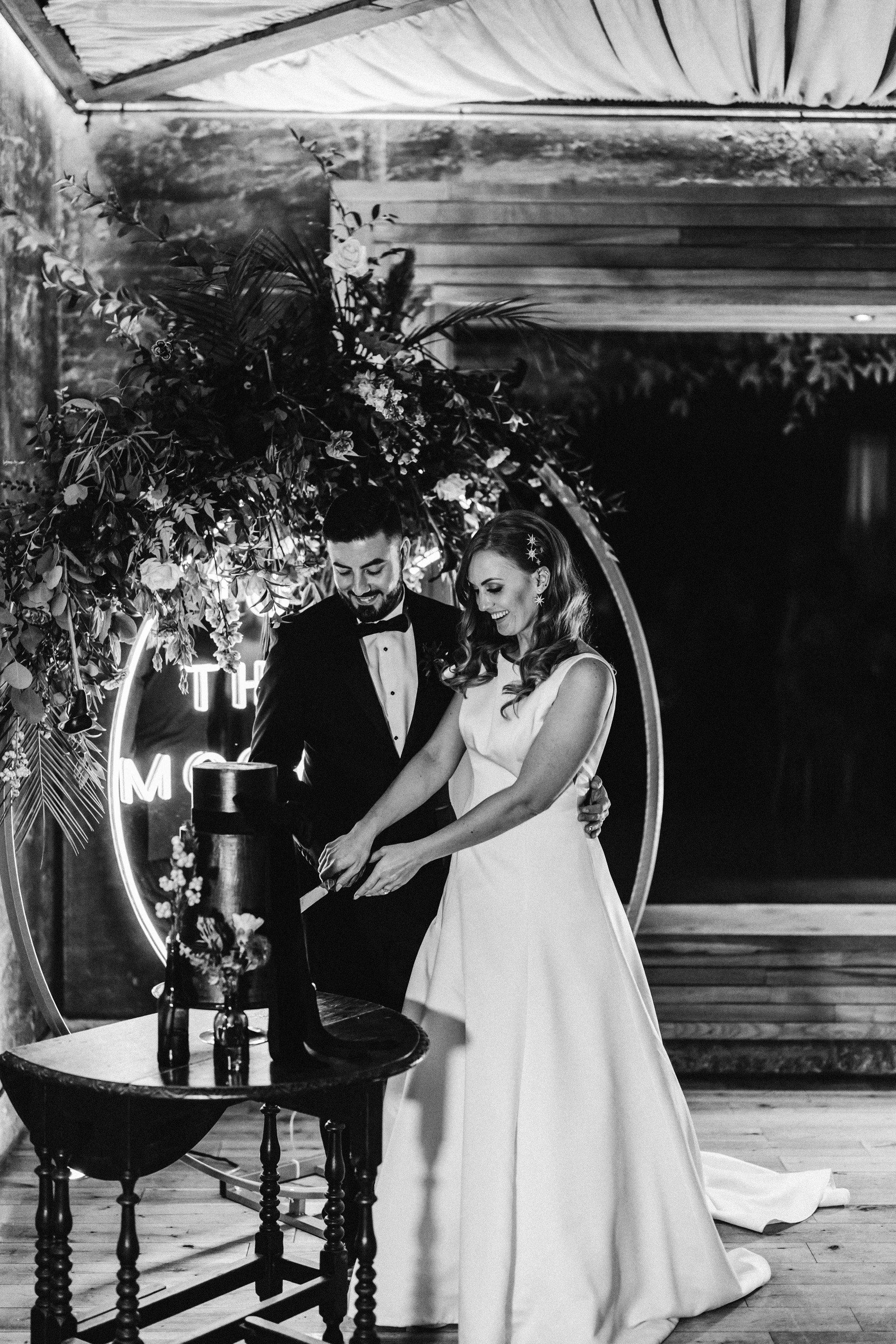 Bride and groom cut their black wedding cake in front of neon moon sign with autumnal flowers at an october wedding with halloween vibes 