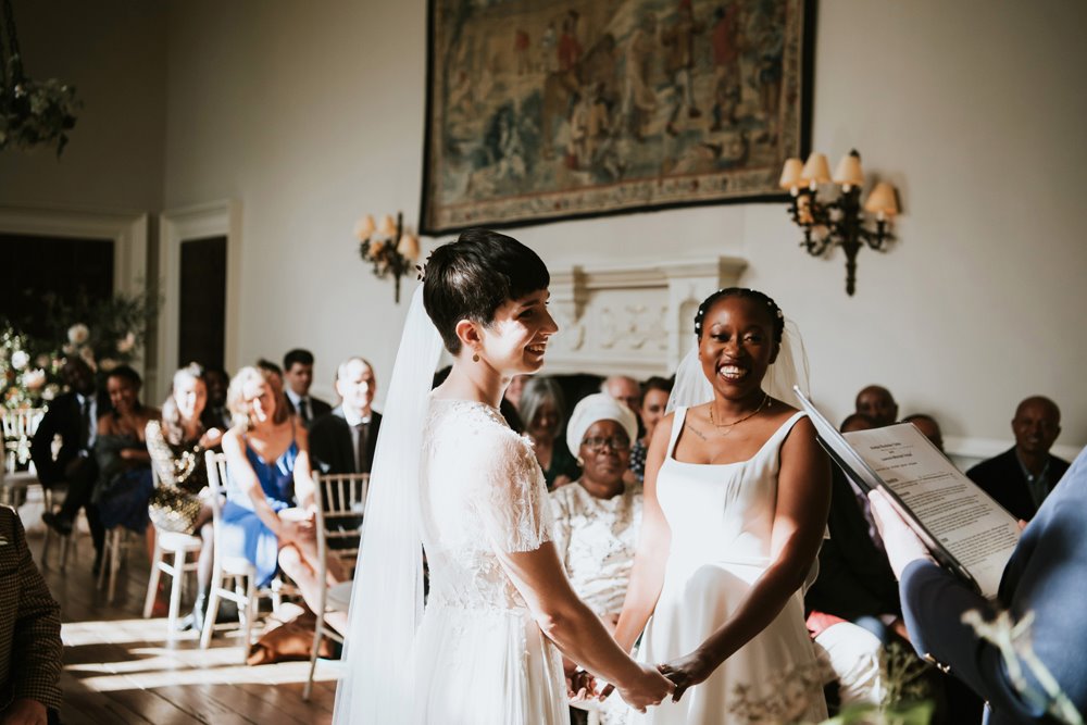 Two modern brides in white wedding dresses and veils hold hands and smile during their same sex wedding ceremony in stately home hall