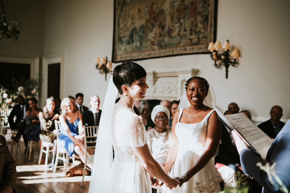 Two modern brides in white wedding dresses and veils hold hands and smile during their same sex wedding ceremony in stately home hall