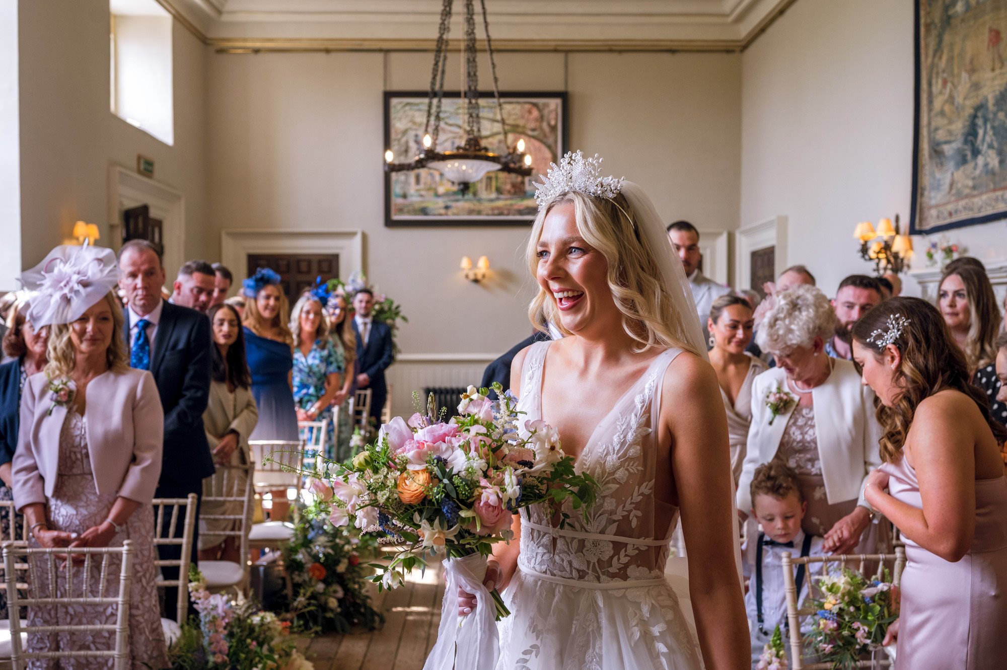 Beautiful blonde boho bride in pearl crown and lace and tulle wedding dress holding bright bouquet at the end of aisle with guests standing behind in historic hall of elmore court