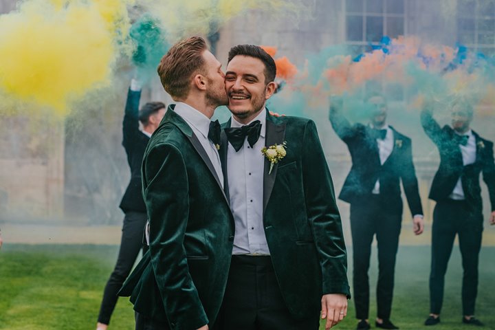 Groom and groom kiss happily on their wedding day with rainbow coloured smoke bombs
