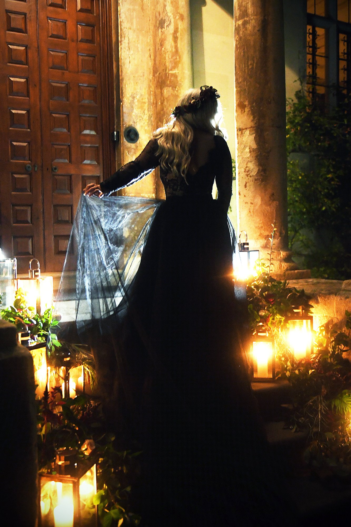 Gothic bride in black wedding dress by candlelight on the steps of her old mansion house wedding venue in england