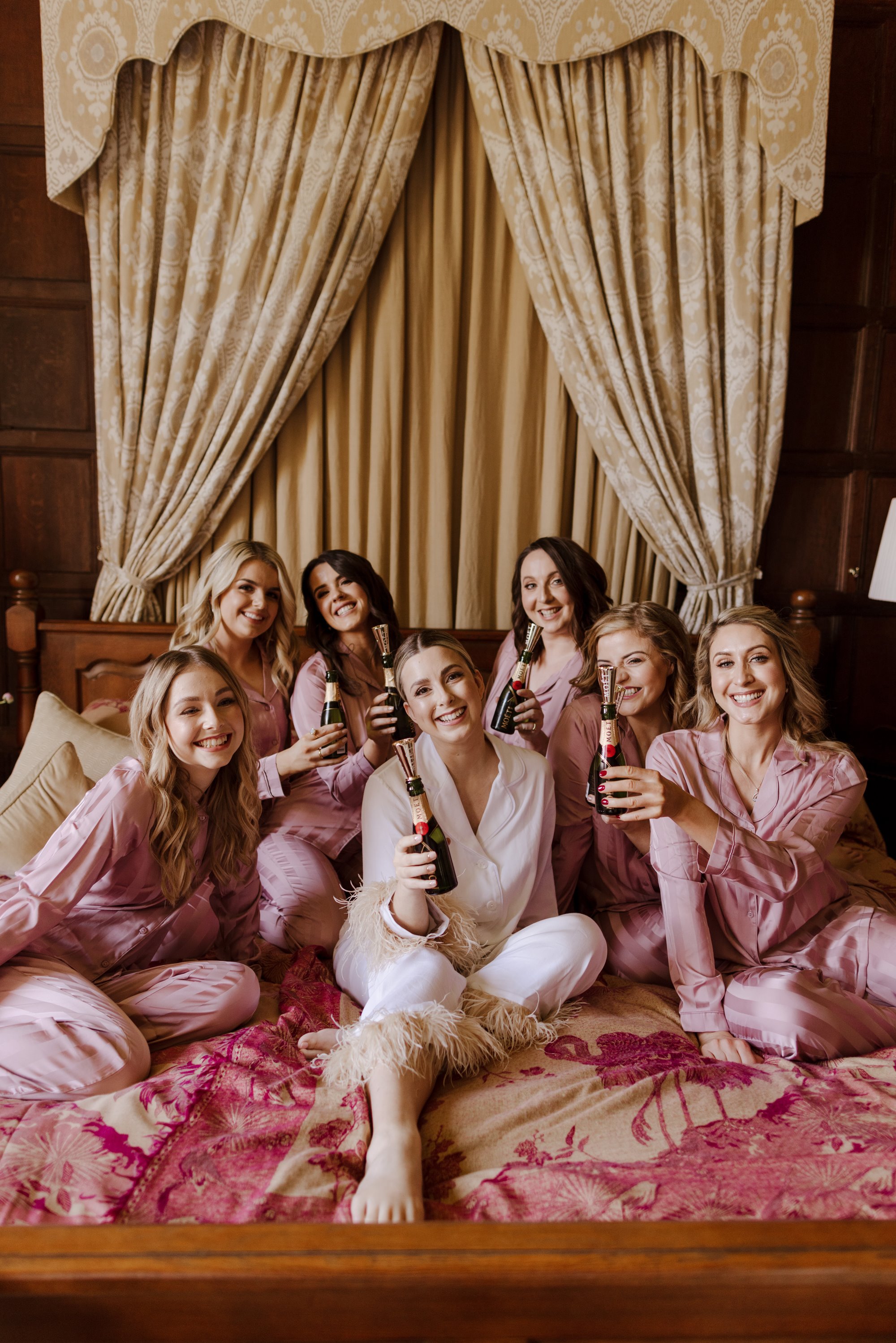 bridal party in pink silk pyjamas, in an elegantly decorated bedroom, toasting with mini bottles of champagne