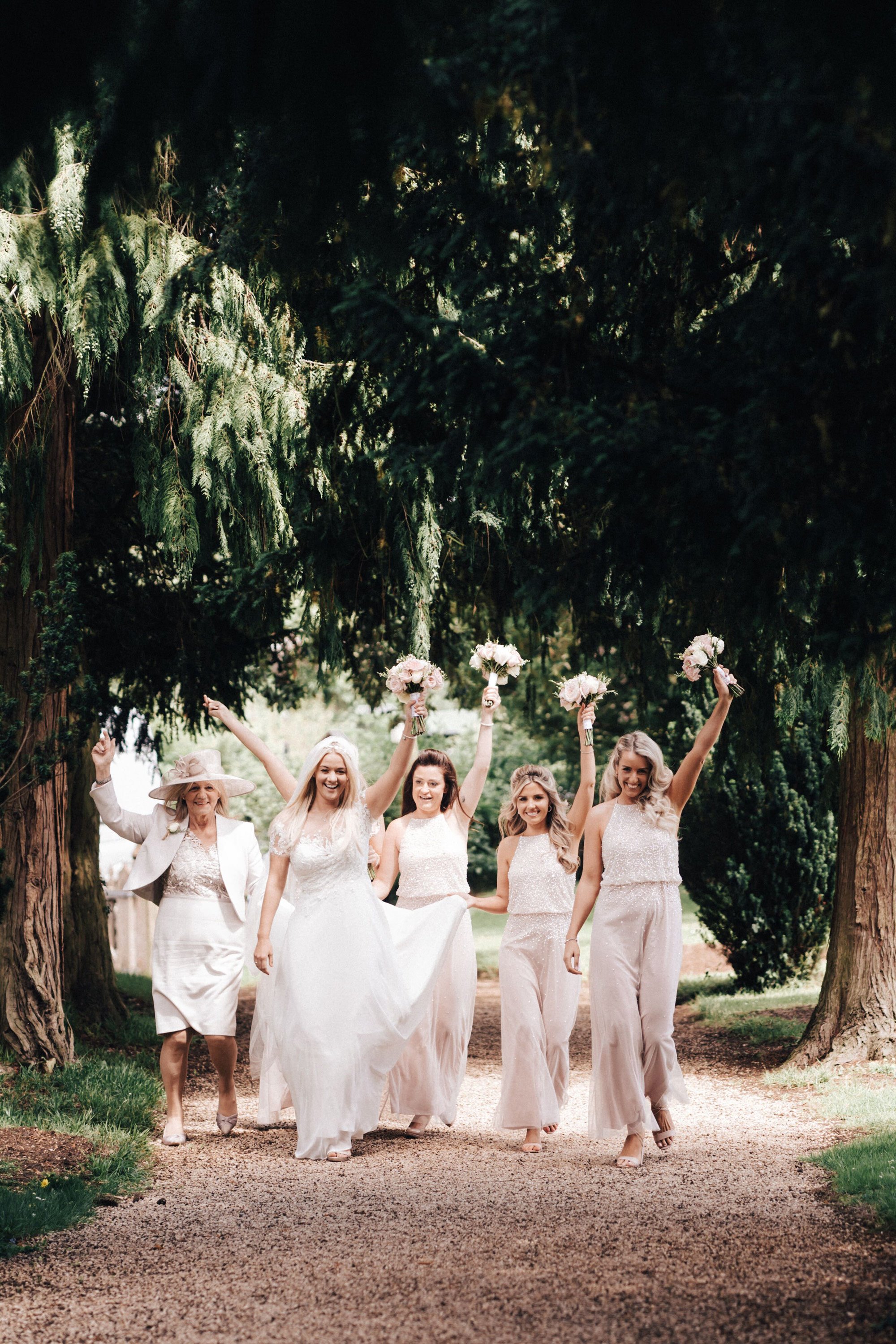 Bridesmaids in pale pink holding bouquets above their heads make their way along the tree lined path to Elmore church for the wedding