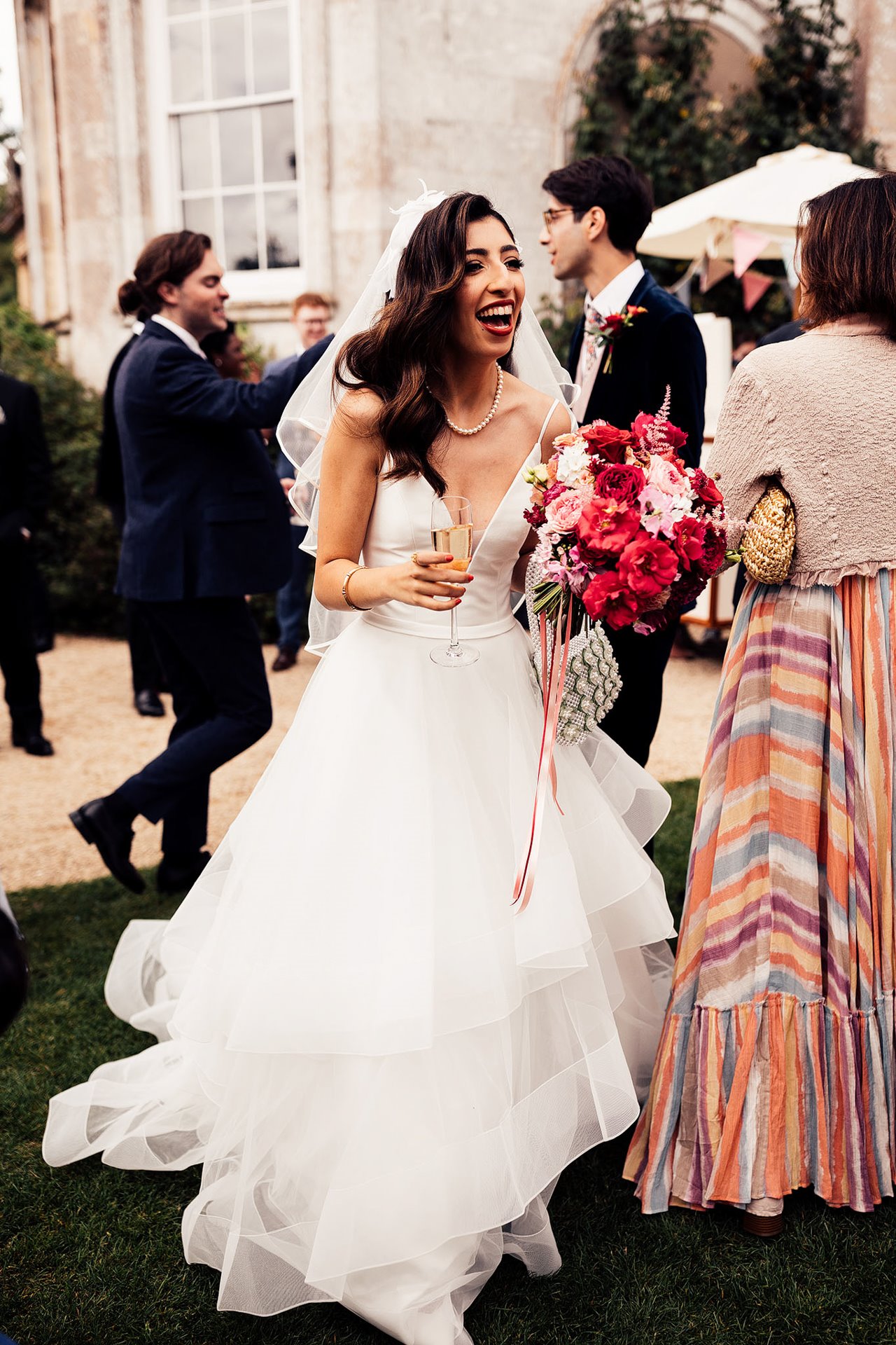 beautiful bride in princess dress holding pink and red bouquet and champagne laughs with guests