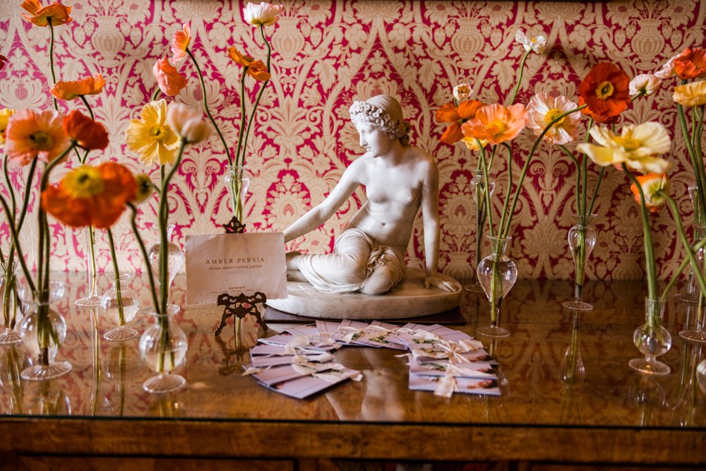 Warm wedding flowers in yellow, orange, red and pink with statue in centre against a red patterned wallpaper at mansion house elmore court in Gloucestershire for a wedding fair