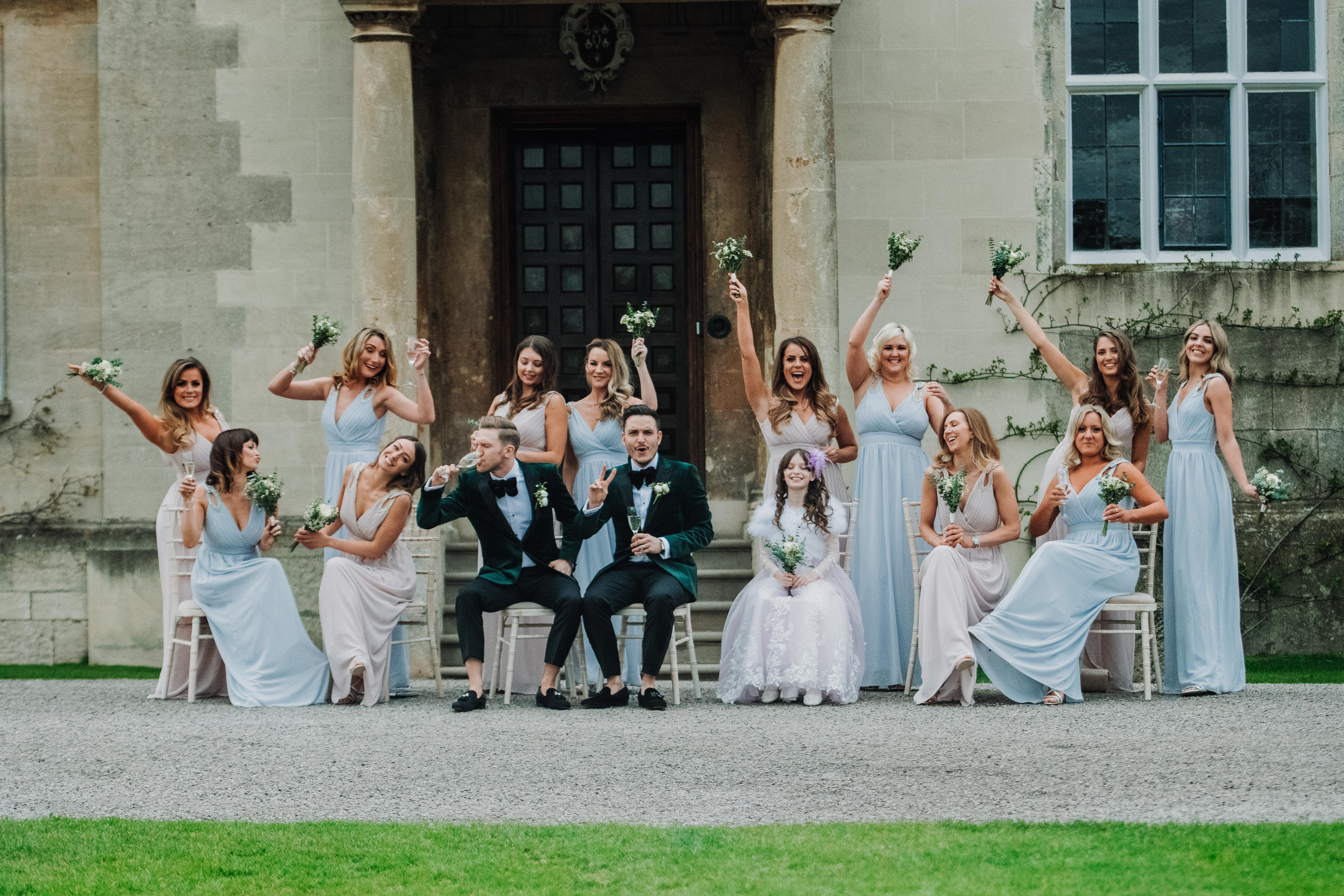 Gay wedding party with 13 bridesmaids having fun posing in front of luxury wedding venue at champagne reception in cotswolds